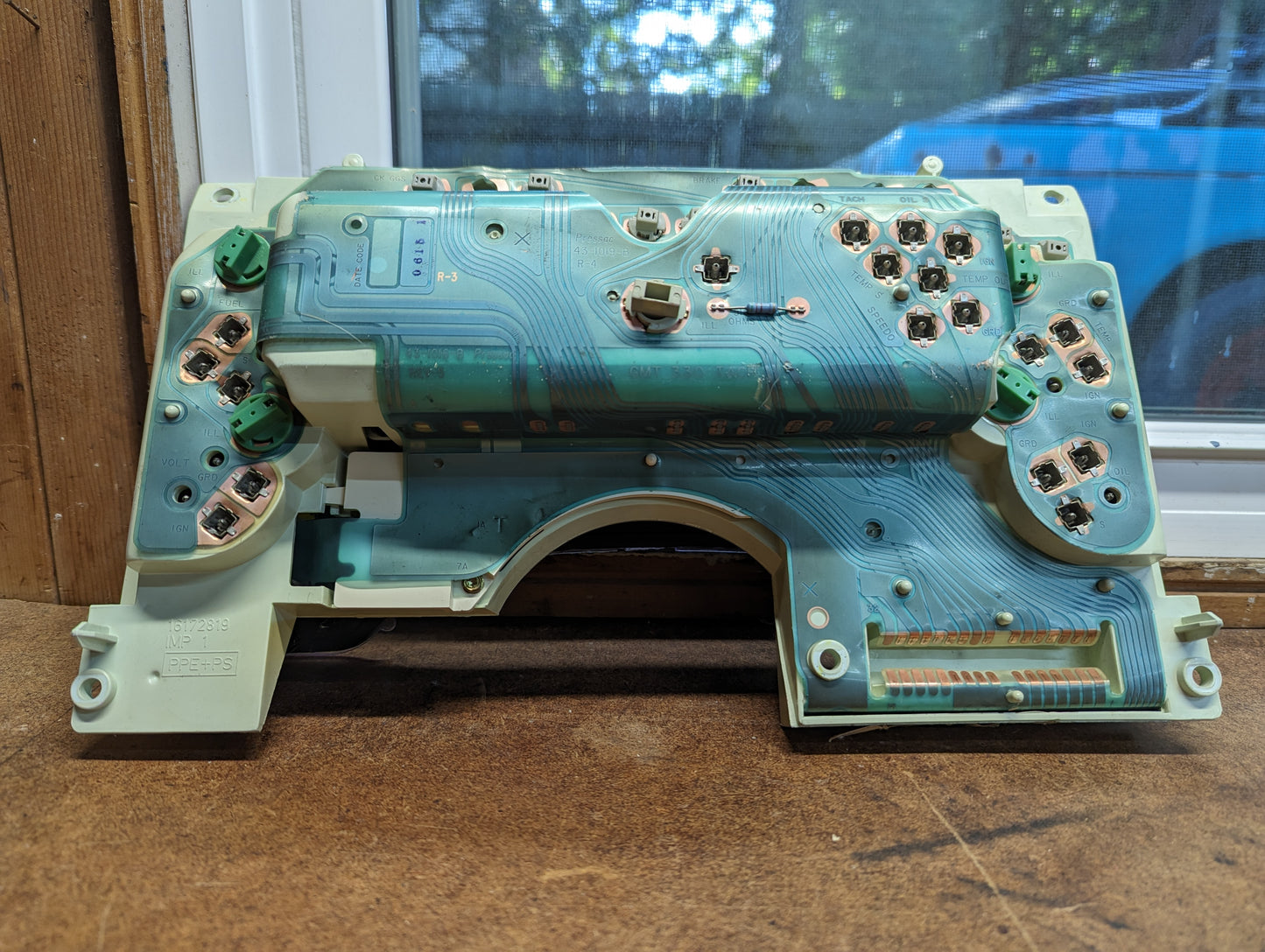 A close-up shows the circuit board of a General Motors OEM Instrument Cluster with Tachometer for 1995-1997 Chevy S10 or Blazer (console shifter), featuring electrical traces, connectors, and bulbs on wood near a window with a blurred vehicle outside.