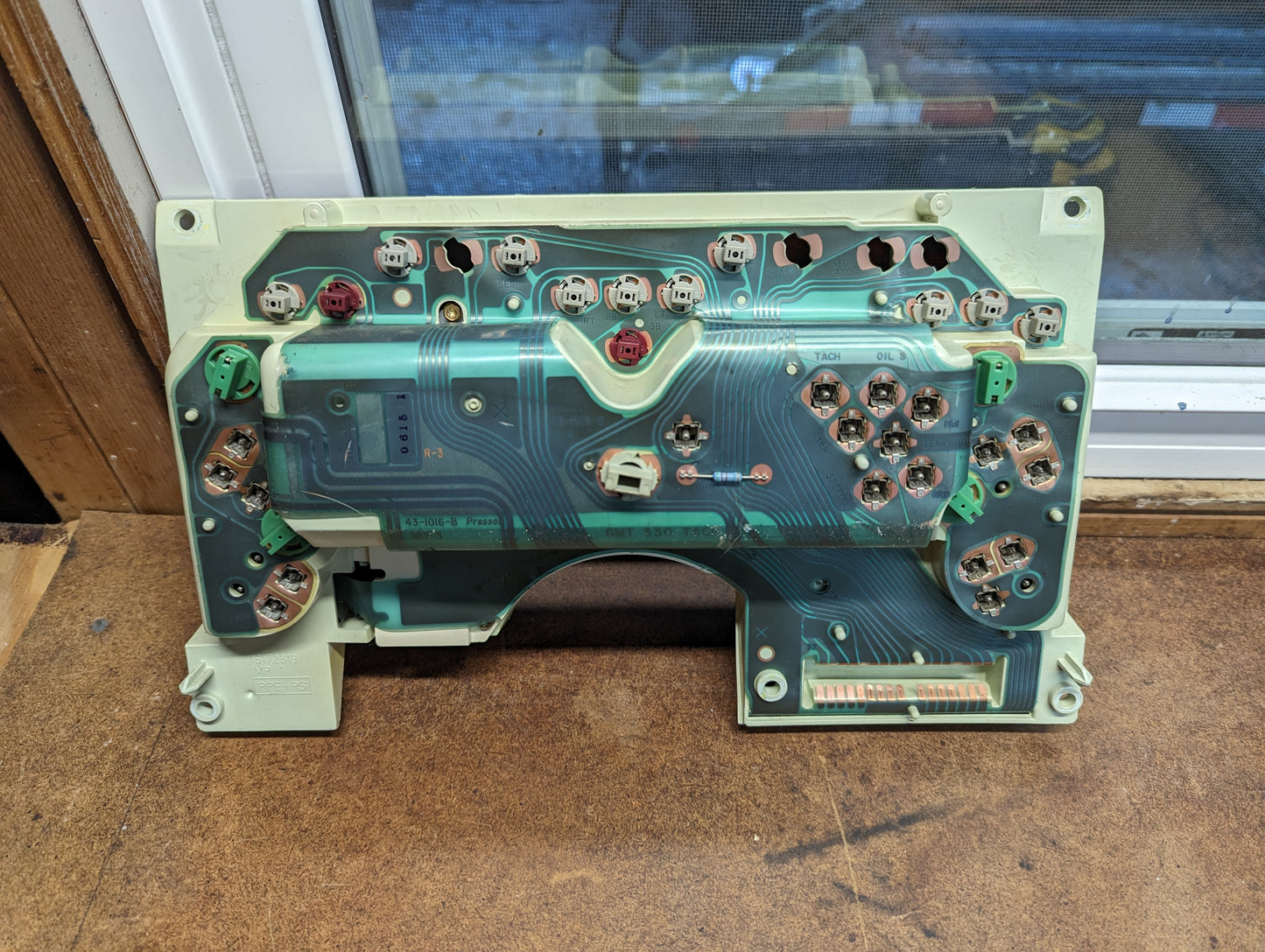 A disassembled General Motors OEM Instrument Cluster with Tachometer for 1995-1997 Chevy S10 or Blazer (console shifter) is shown on a wooden surface, displaying its connectors and copper traces.