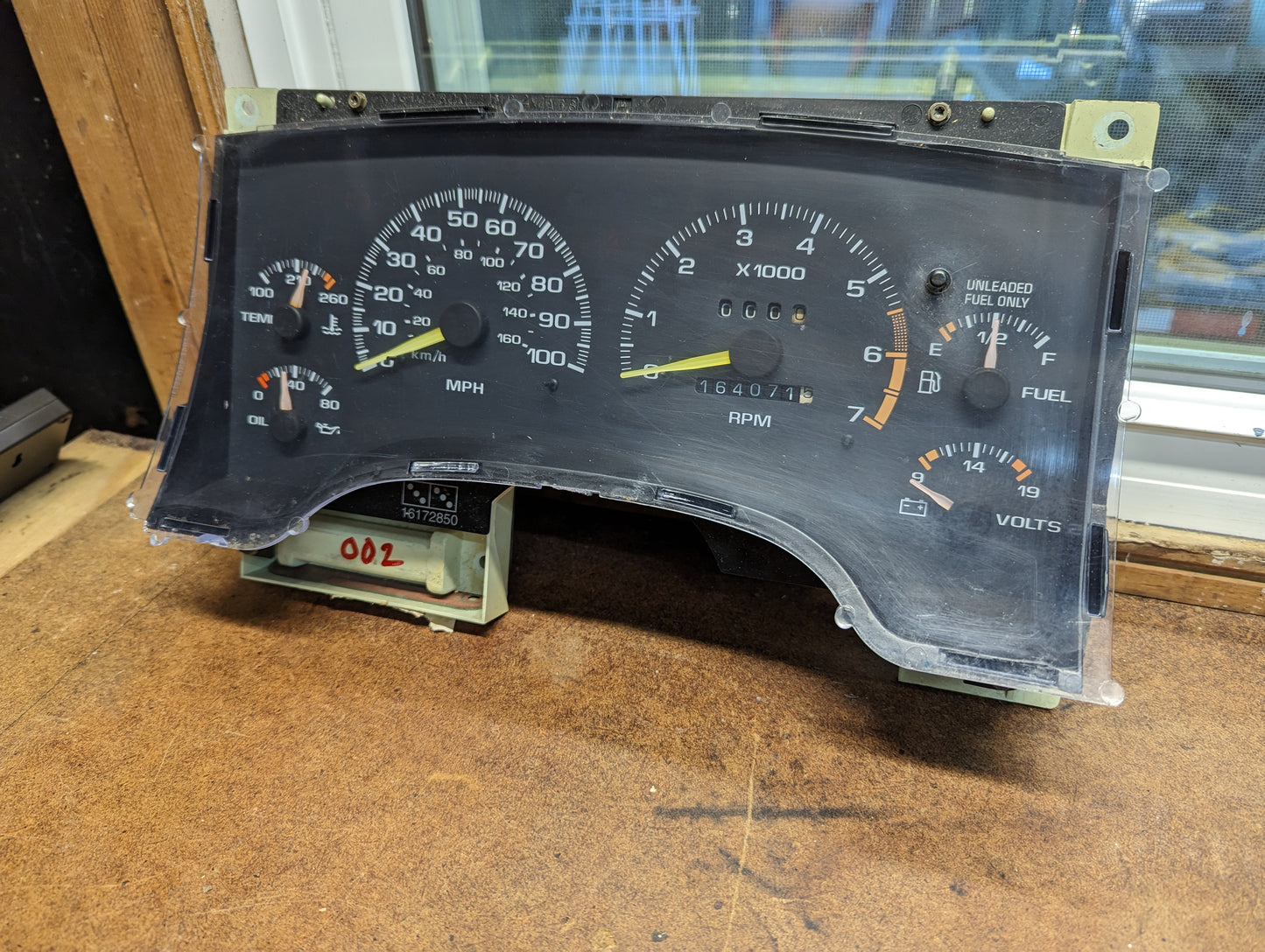 A General Motors OEM Instrument Cluster with Tachometer for 1995-1997 Chevy S10 or Blazer with Console Shifter rests on a wooden surface near a window.