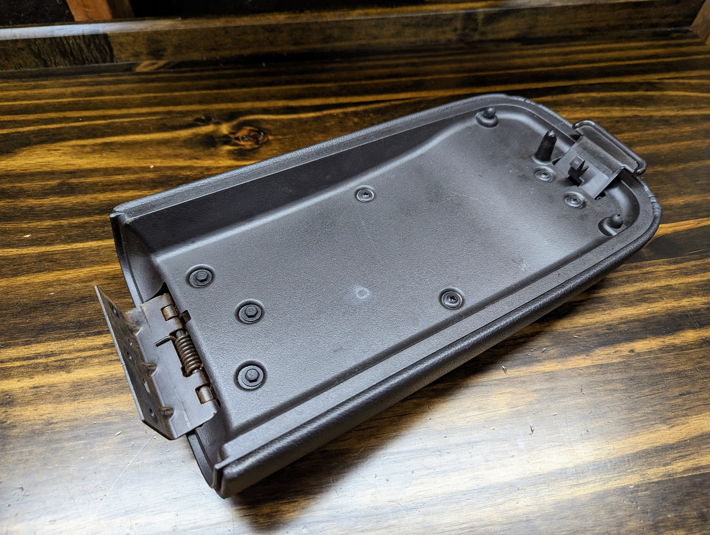 A General Motors Leather Armrest Center Console Lid in black for 2002-2009 GMC Envoy and Chevy Trailblazer is lying on a wooden surface, shown from the underside.