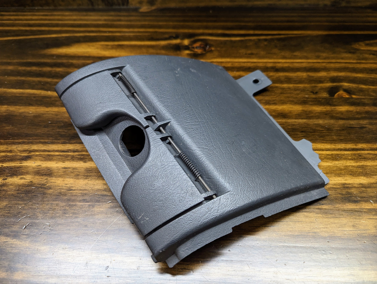 A General Motors OEM 3 Port 12V Aux Panel Ash Tray Delete in dark gray for 1998-2000 GMC Envoy, shown on a wooden surface, features textured finishes, a small side mounting tab, and complements charcoal interior panels.