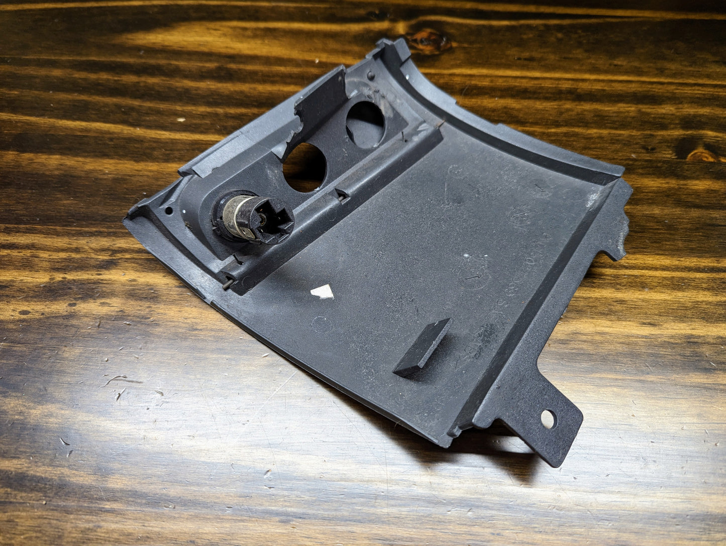 A dark gray, rectangular OEM 3 Port 12V Aux Panel Ash Tray Delete by General Motors for 1998–2000 GMC Envoy rests on a dark wooden surface. It has cutouts, angled edges, mounting tabs, and shows signs of wear.