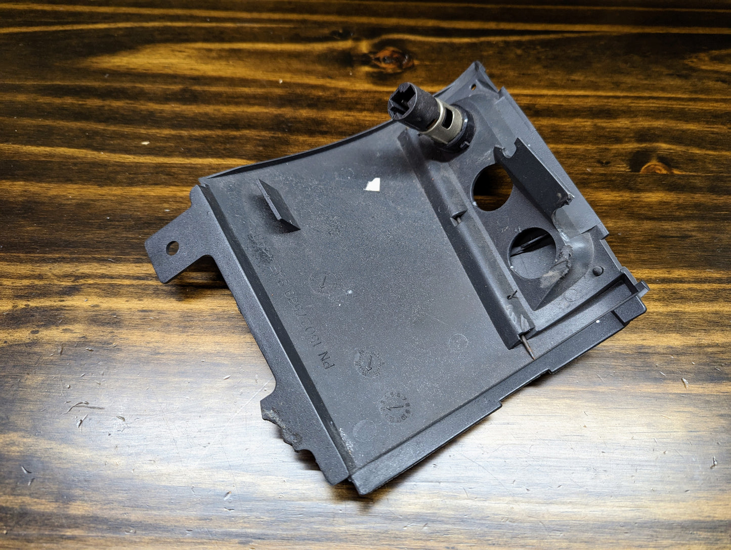 A dark gray OEM 3 Port 12V Aux Panel Ash Tray Delete by General Motors, from a 1998–2000 GMC Envoy, with sensor attached and mounting holes, is shown slightly worn on a wooden surface.