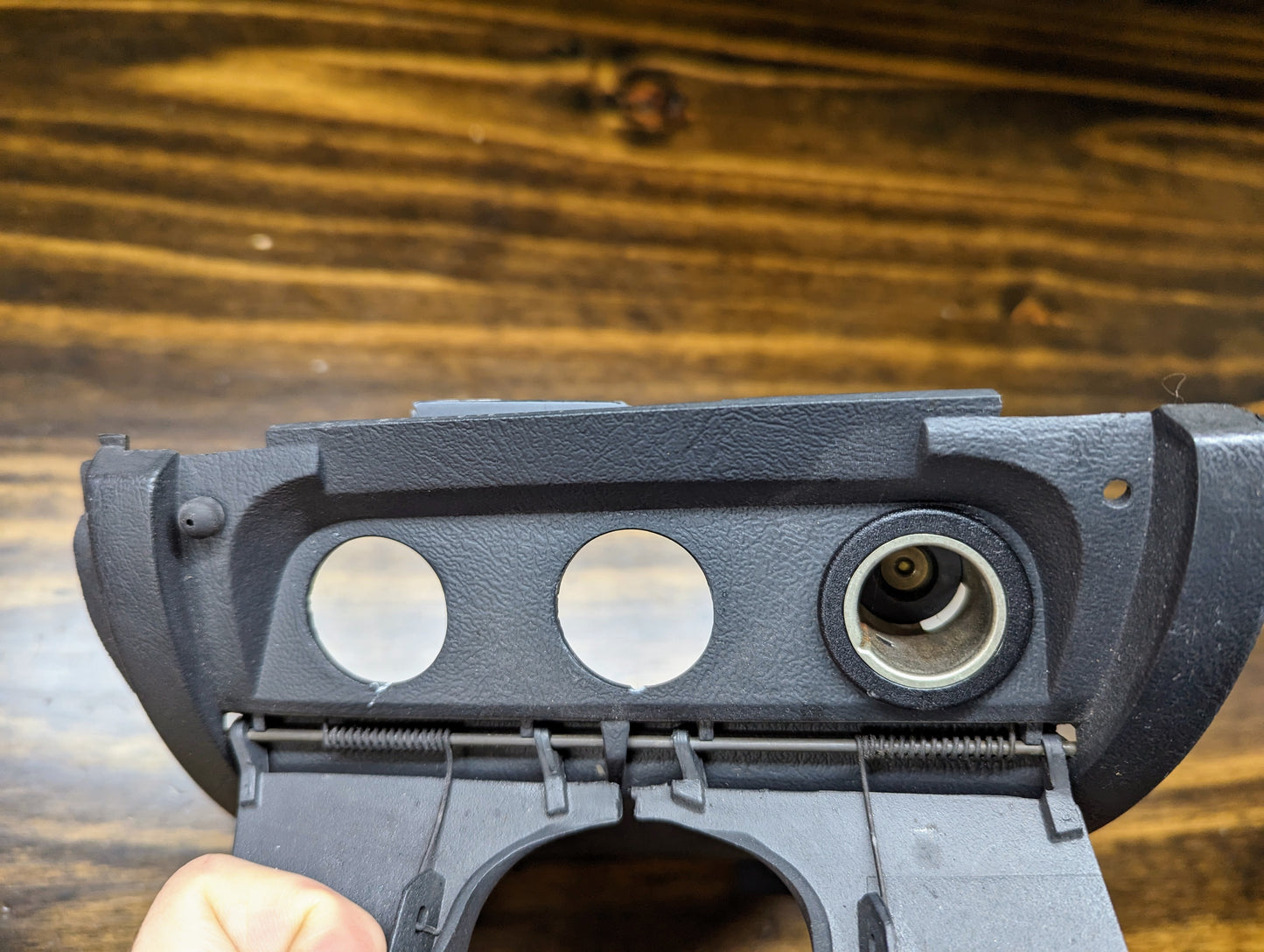 A hand holds a General Motors OEM 3 Port 12V Aux Panel Ash Tray Delete in Dark Gray for 1998-2000 GMC Envoy, featuring two circular holes and a cigarette lighter socket, shown close-up against a wooden background.