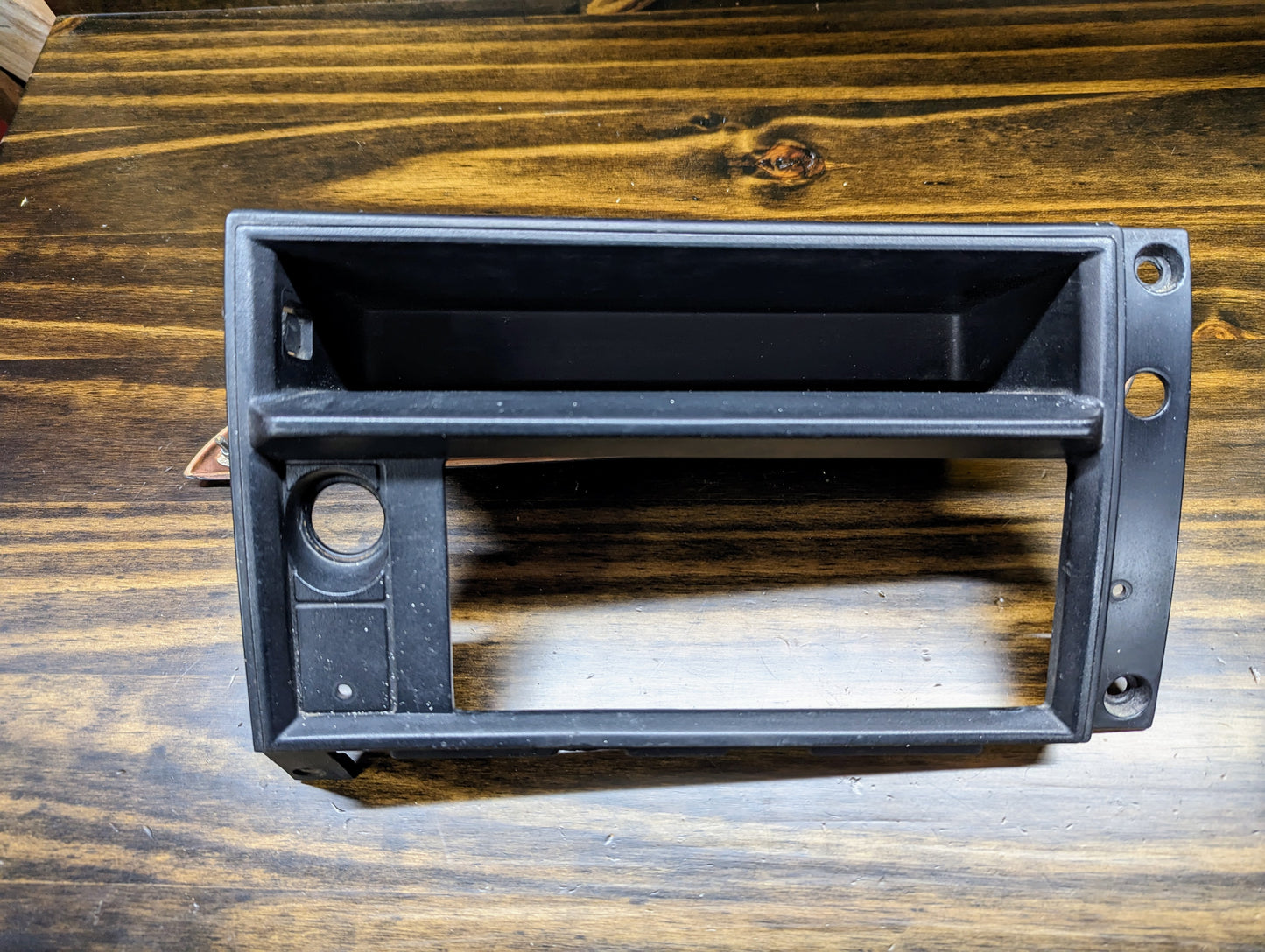 A General Motors OEM Radio Dash Bezel w/Vent Delete in Black for 1986-1993 Chevy S10 Blazer, GMC S15, and more is shown on a wooden surface, featuring a rectangular opening and small circular hole.
