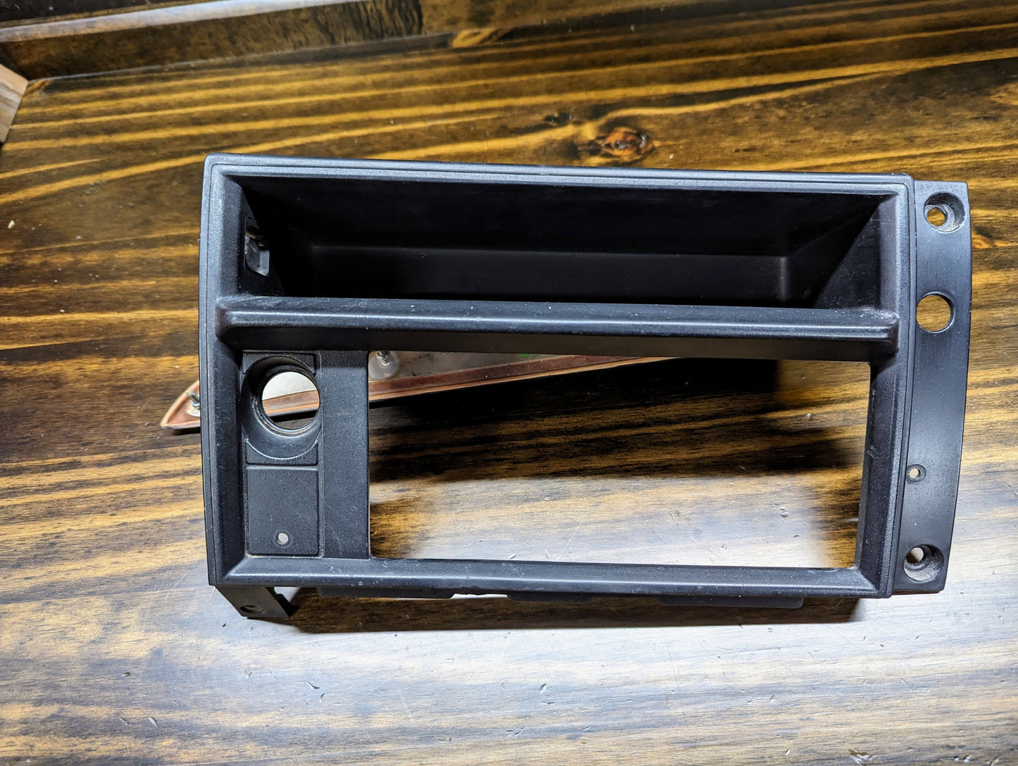 A black OEM Radio Dash Bezel w/Vent Delete by General Motors for 1986-1993 Chevy S10 Blazer, GMC S15, and more, featuring two rectangular slots, a circular hole, and two right-side screw holes on a dark wood surface.