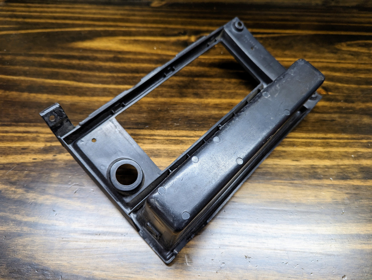 A General Motors OEM Radio Dash Bezel w/Vent Delete in black for 1986-1993 Chevy S10, Blazer, GMC S15, and more, featuring a rectangular opening and rounded edges, shown on a dark streaked wooden surface.