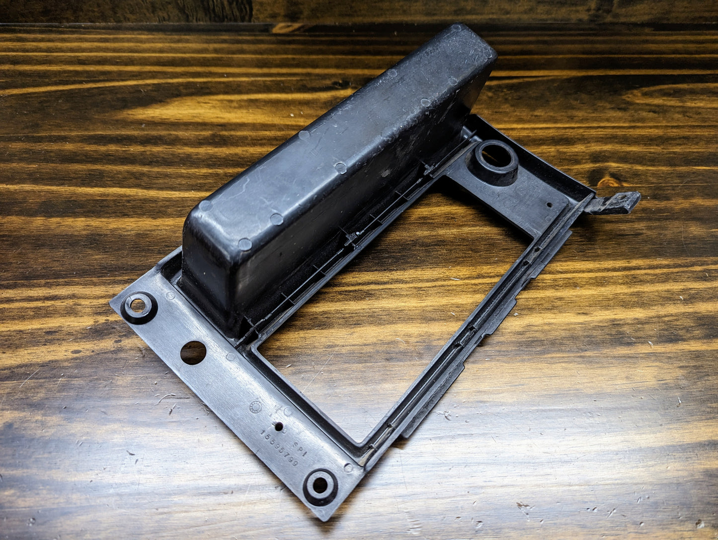 A black OEM Radio Dash Bezel w/Vent Delete by General Motors for 1986-1993 Chevy S10, Blazer, GMC S15, and more, with a rectangular opening and mounting holes, shows light wear from prior use and is displayed on a wood-grain surface.