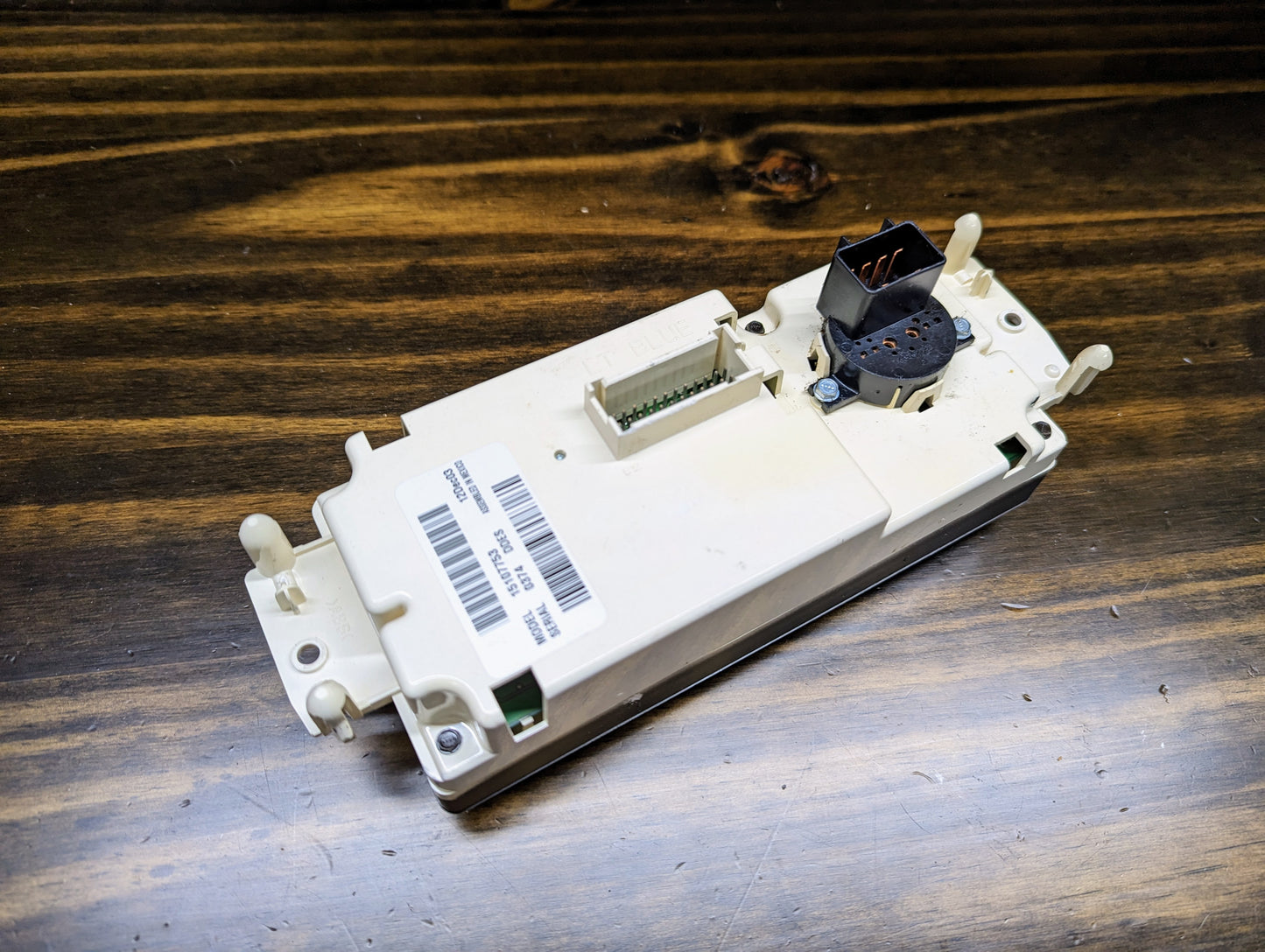A beige General Motors OEM Dual Climate HVAC Manual Control Head with Rear Defrost for 2003-2006 GM Full Size Trucks, featuring multiple connectors and a barcode label, is shown on a wooden surface with visible copper wires on a connector.