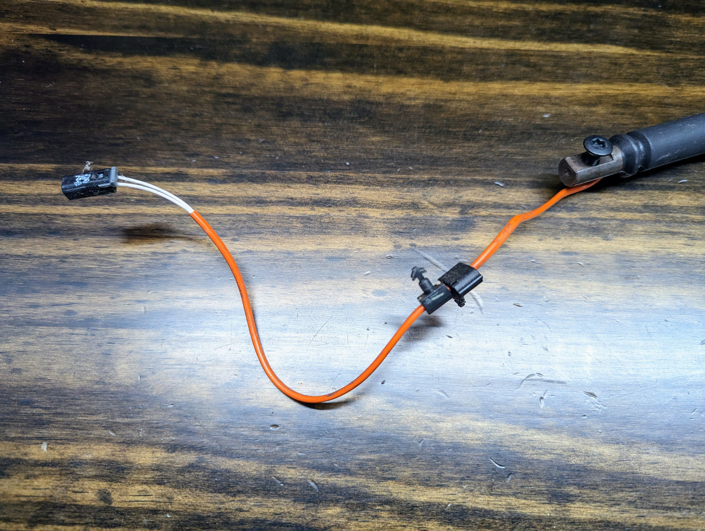 An orange and white wire with a black connector and sensor is shown near a General Motors OEM Column Shift Lever with Gray Leather Wrap for 1999-2006 GM Full Size Trucks, resting on wood with a black cylindrical part to the right.