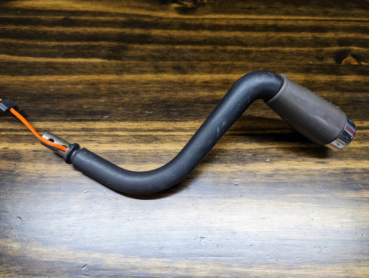 A black, S-shaped General Motors OEM Column Shift Lever with Gray Leather Wrap for 1999-2006 GM Full Size Trucks, featuring an orange wire and metallic tip, rests on a dark wooden surface.