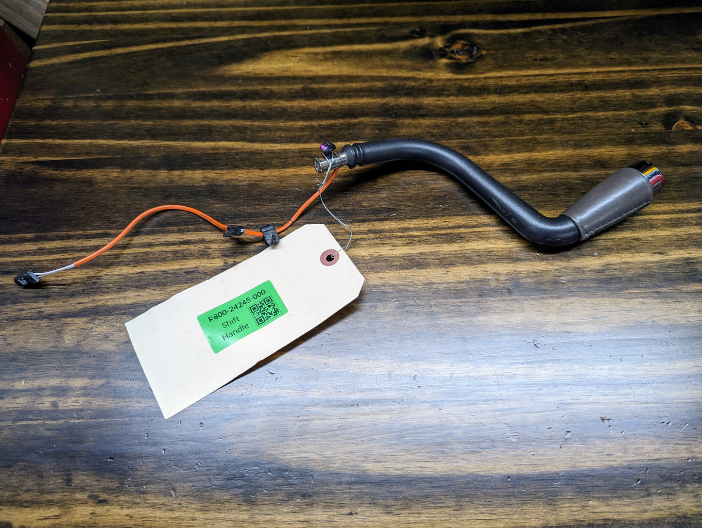 A curved black OEM Column Shift Lever with gray leather wrap for 1999-2006 GM Full Size Trucks by General Motors, featuring a multicolored connector and paper tag labeled P/N: 3244-600 Static Harness, rests on a dark wooden surface.