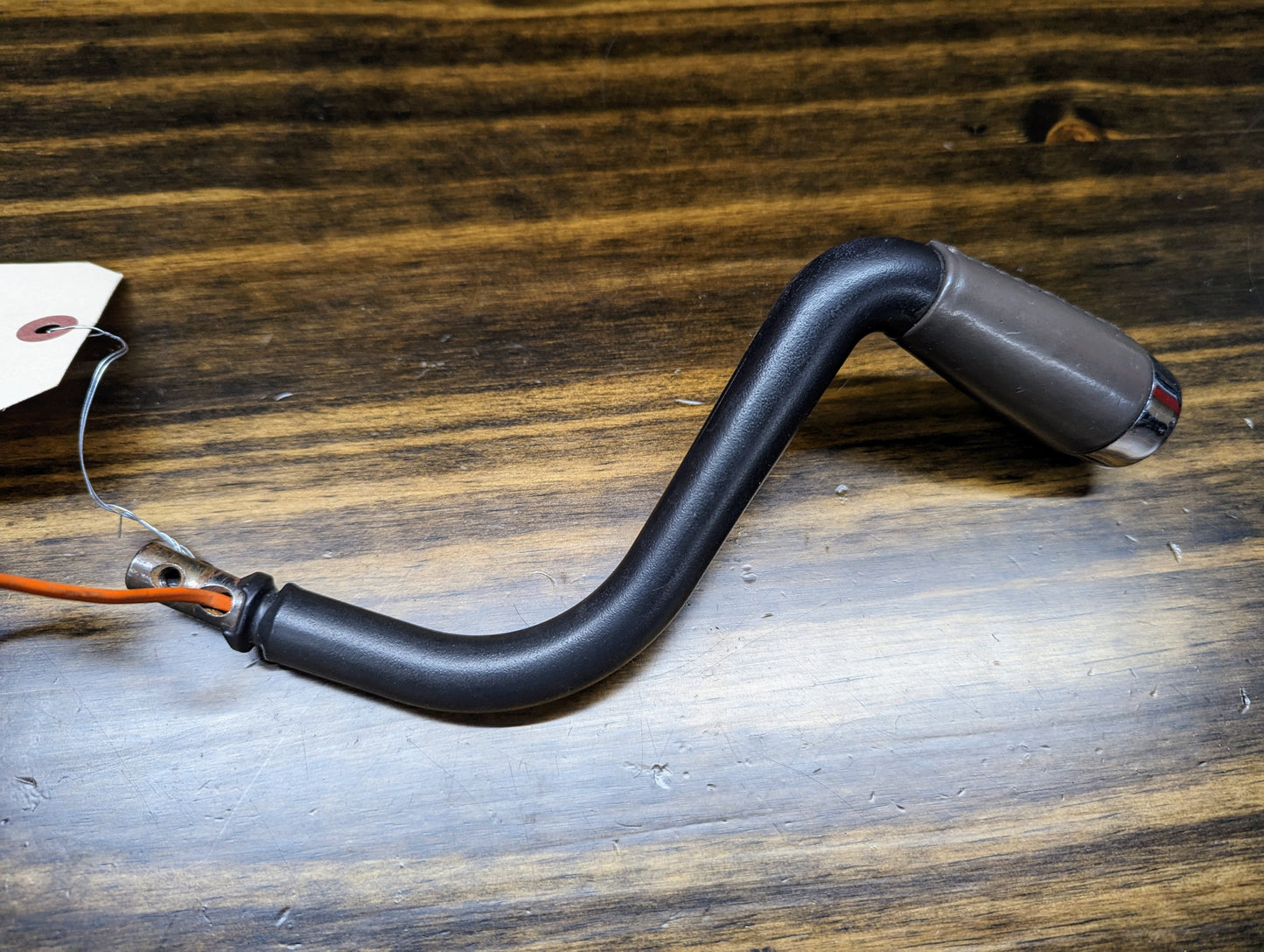 A General Motors OEM column shift lever with gray leather wrap for 1999-2006 GM full size trucks, featuring an L-shaped black handle and attached orange wire with tag, is displayed on a wooden surface.