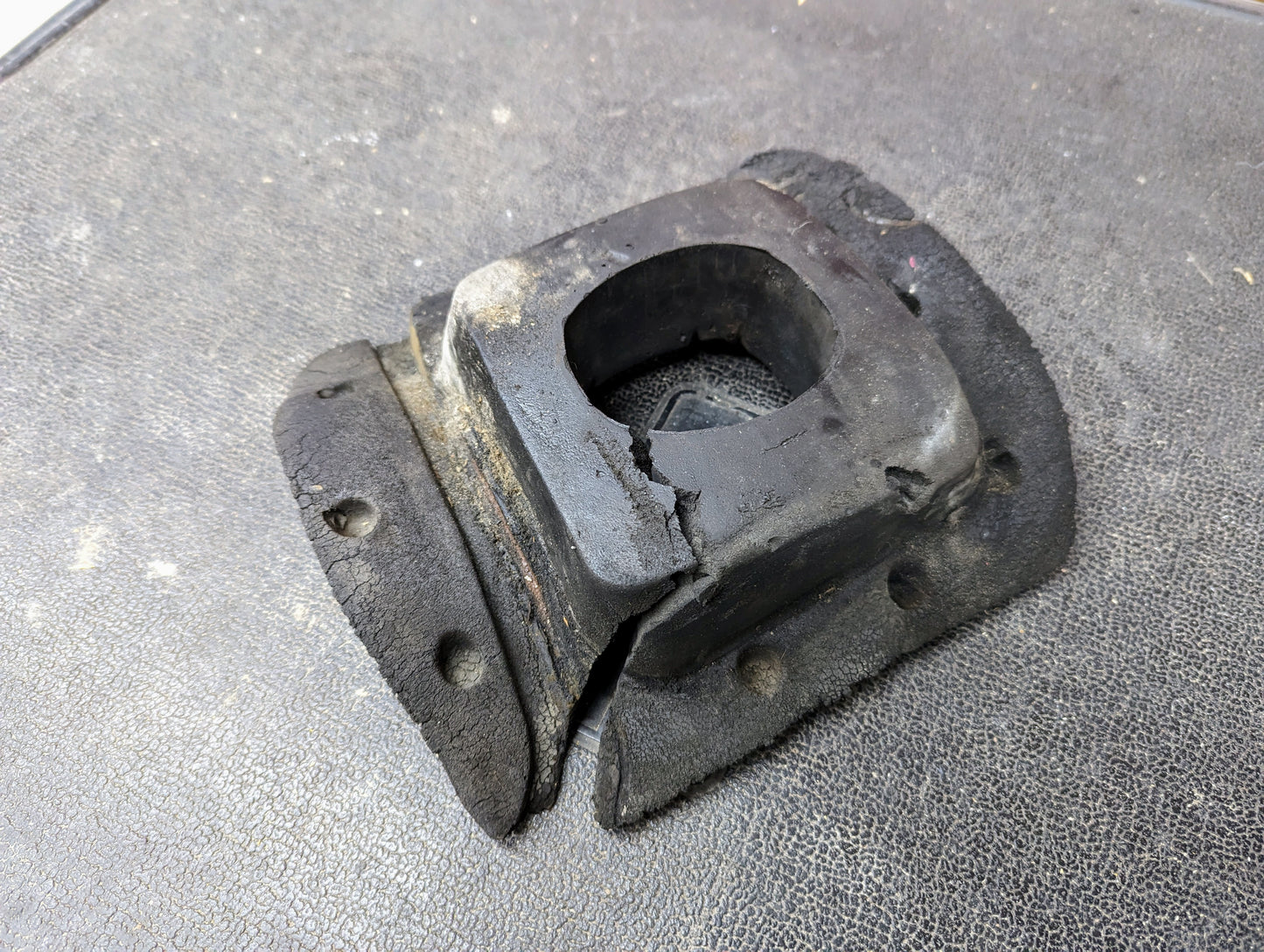 A worn and cracked General Motors Manual Floor Shifter Insulator Foam for 1998-2005 Chevy S10, Blazer, GMC Sonoma, or Jimmy shows significant wear, with splits and bolt holes visible on the textured black rubber surface.