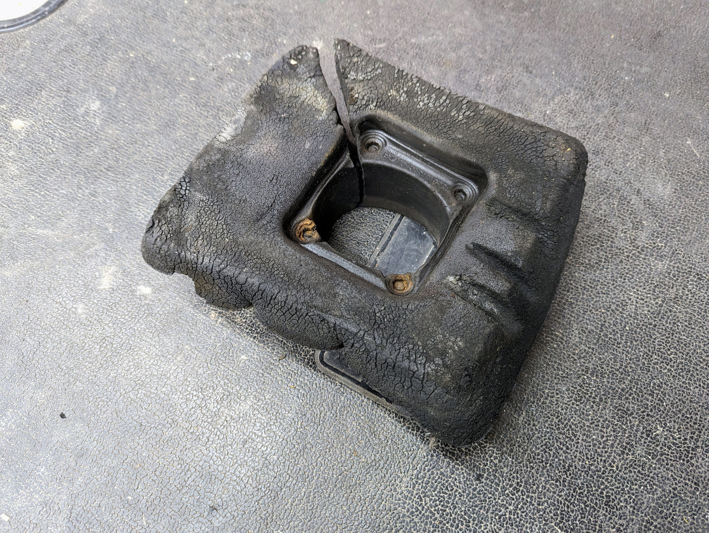 A worn General Motors Manual Floor Shifter Insulator Foam for 1998-2005 Chevy S10 Blazer GMC Sonoma Jimmy lies on a textured dark surface. Four screws hold the center, with visible cracks and a partly detached rubber strip.
