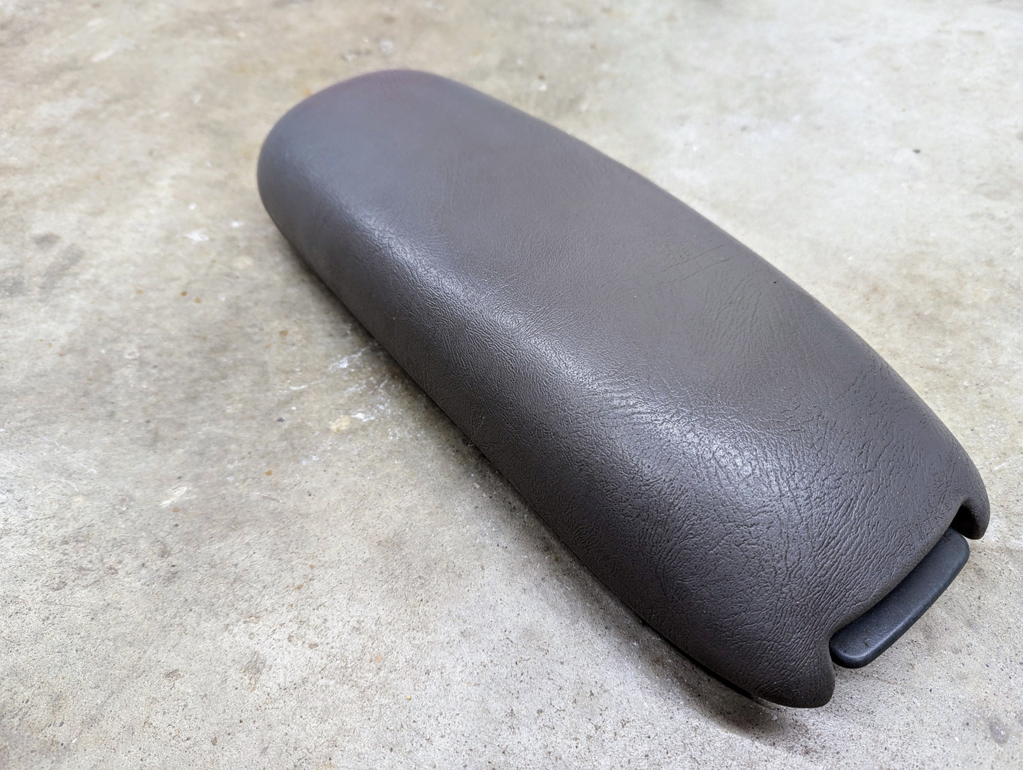A General Motors OEM Center Console Lid Armrest in light gray for 1998-2005 Chevy S10, Blazer, and more is shown on a concrete surface with a textured finish and latch or hinge on one end.