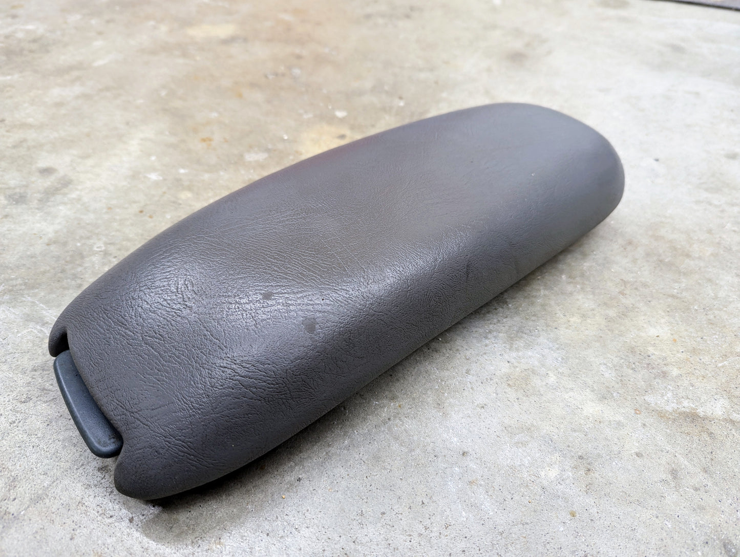 A General Motors OEM Center Console Lid Armrest in light gray for 1998-2005 Chevy S10 and Blazer rests on concrete, displaying a padded, slightly textured finish with a hinge mechanism on one end.