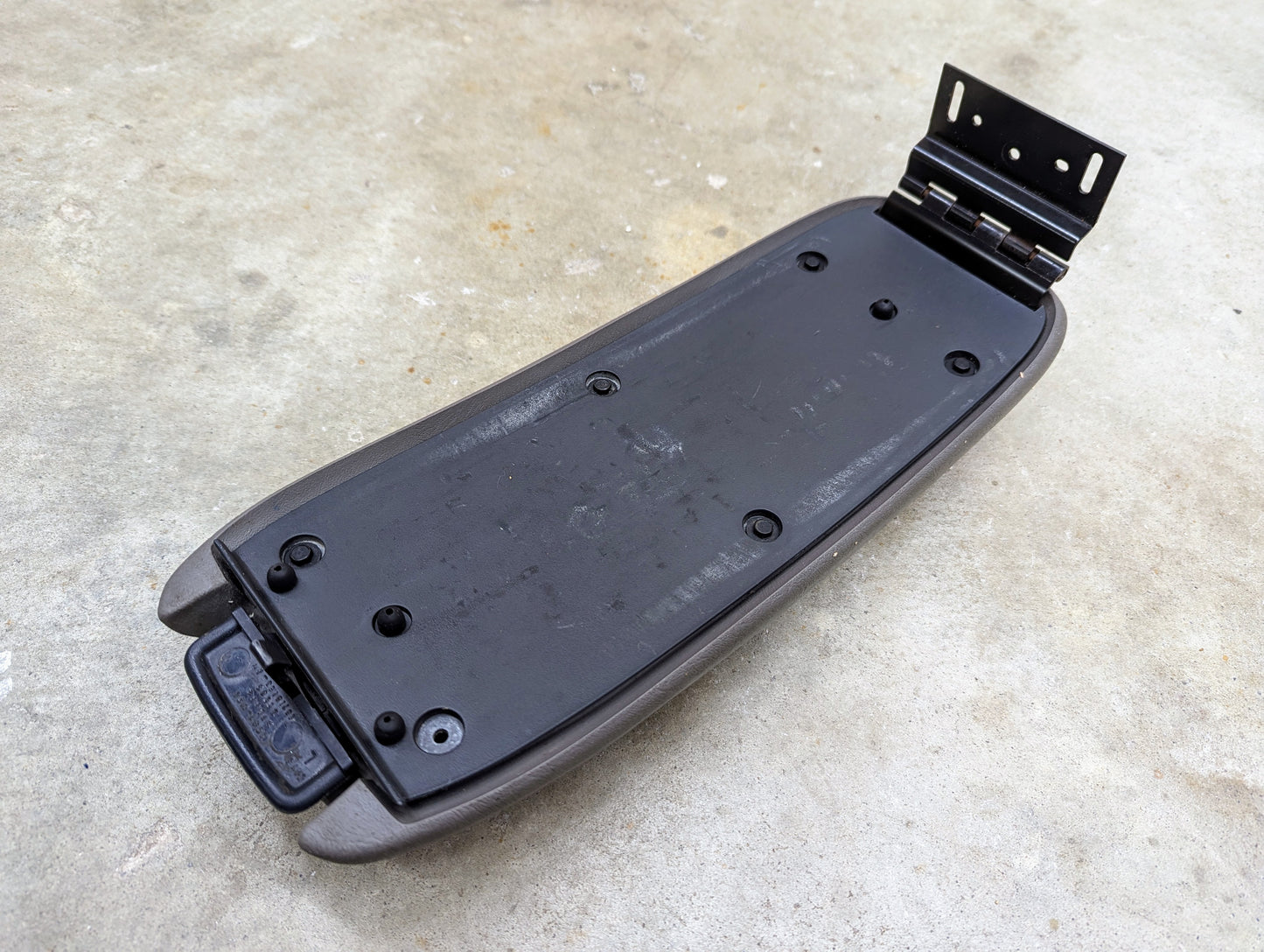 A Light Gray OEM Center Console Lid Armrest by General Motors for 1998-2005 Chevy S10, Blazer, and more, with hinge hardware attached, is shown upside down on a concrete surface.