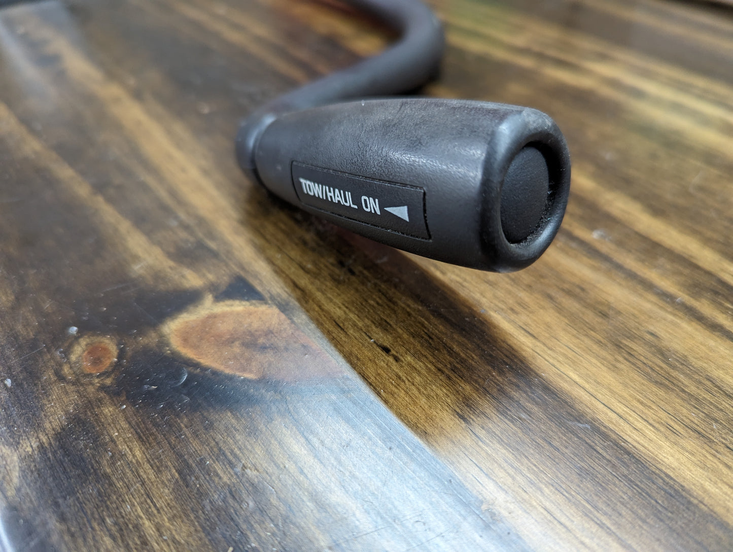 A close-up of the General Motors OEM Column Shift Lever with Tow/Haul for 1999-2006 GM Full Size Trucks, featuring the TOW/HAUL ON label and a slightly curved handle, shown resting on a wooden surface.