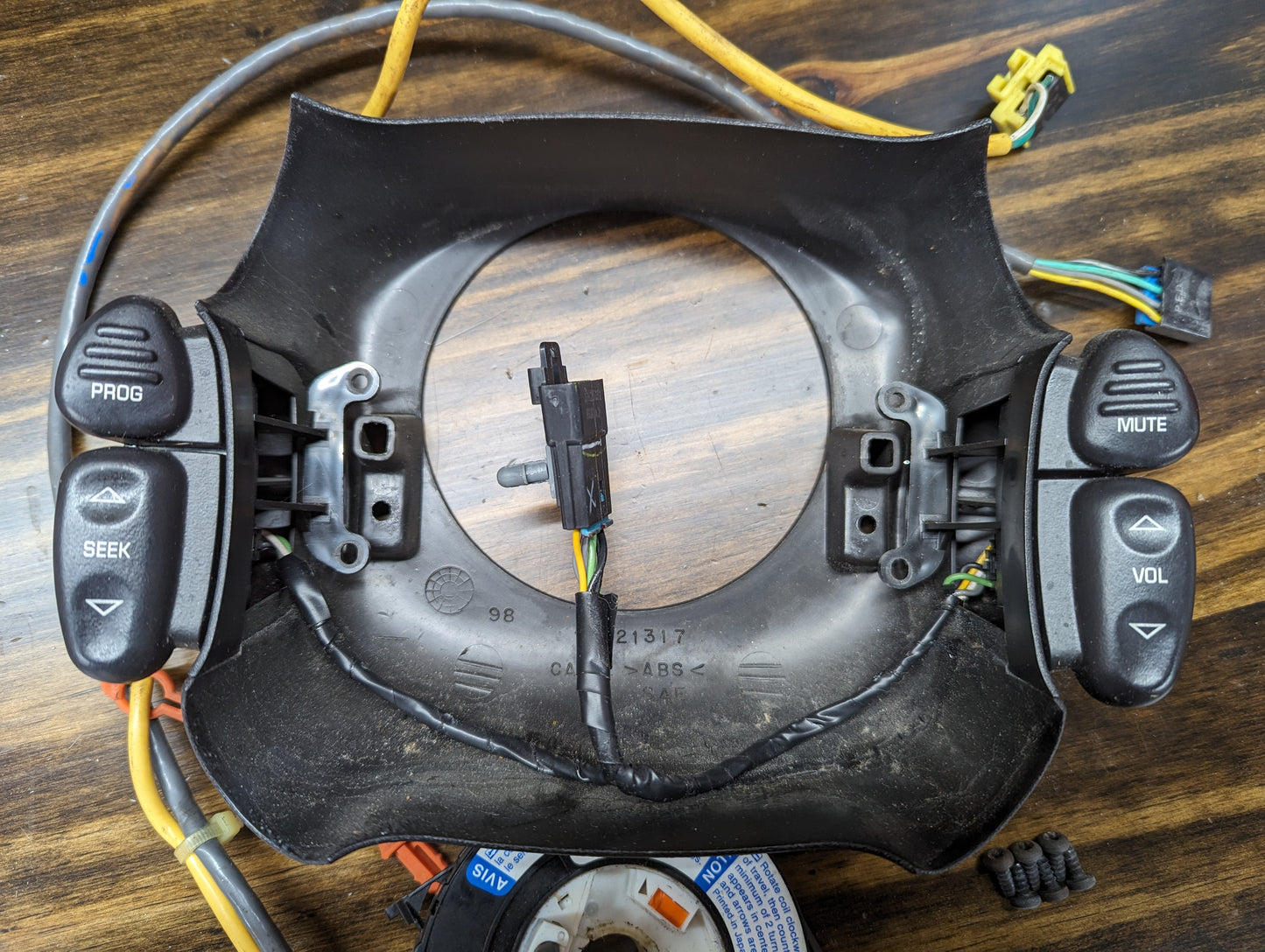 A General Motors KIT Steering Wheel Black Button Radio Controls & Clock Spring for 2000-2003 Chevy S10, Blazer, and more lies face down on a wooden surface, showing its attached wiring and connectors.