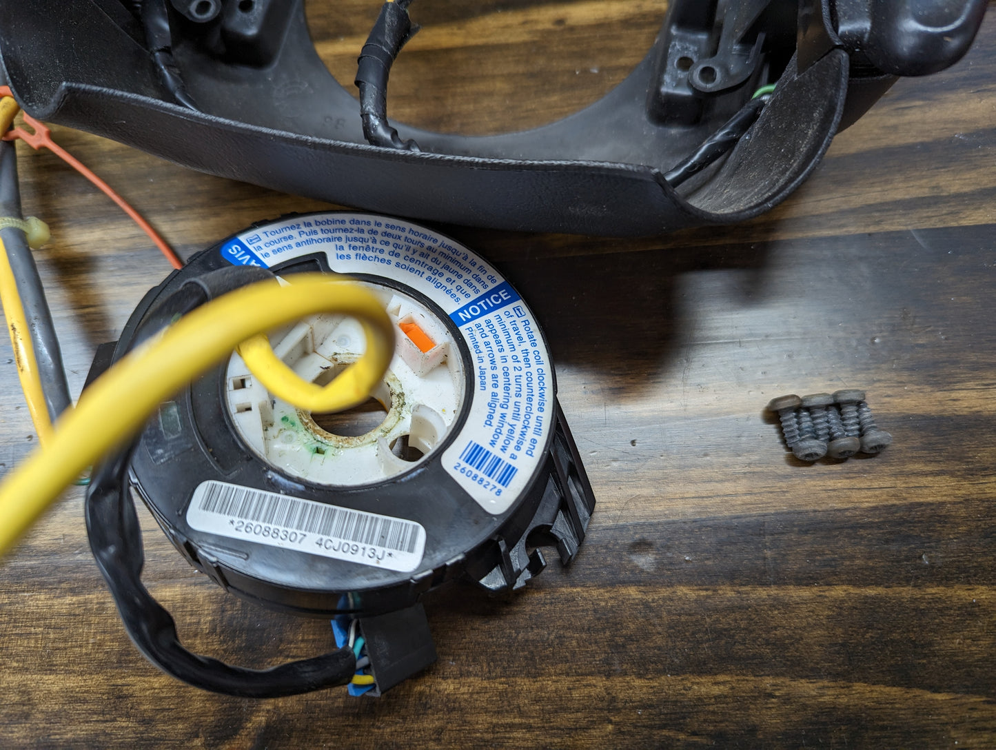 A disassembled General Motors KIT Steering Wheel Black Button Radio Controls & Clock Spring for 2000-2003 Chevy S10, Blazer, and more rests on wood with a yellow cable and screws. The circular part displays labels, numbers, and warnings.
