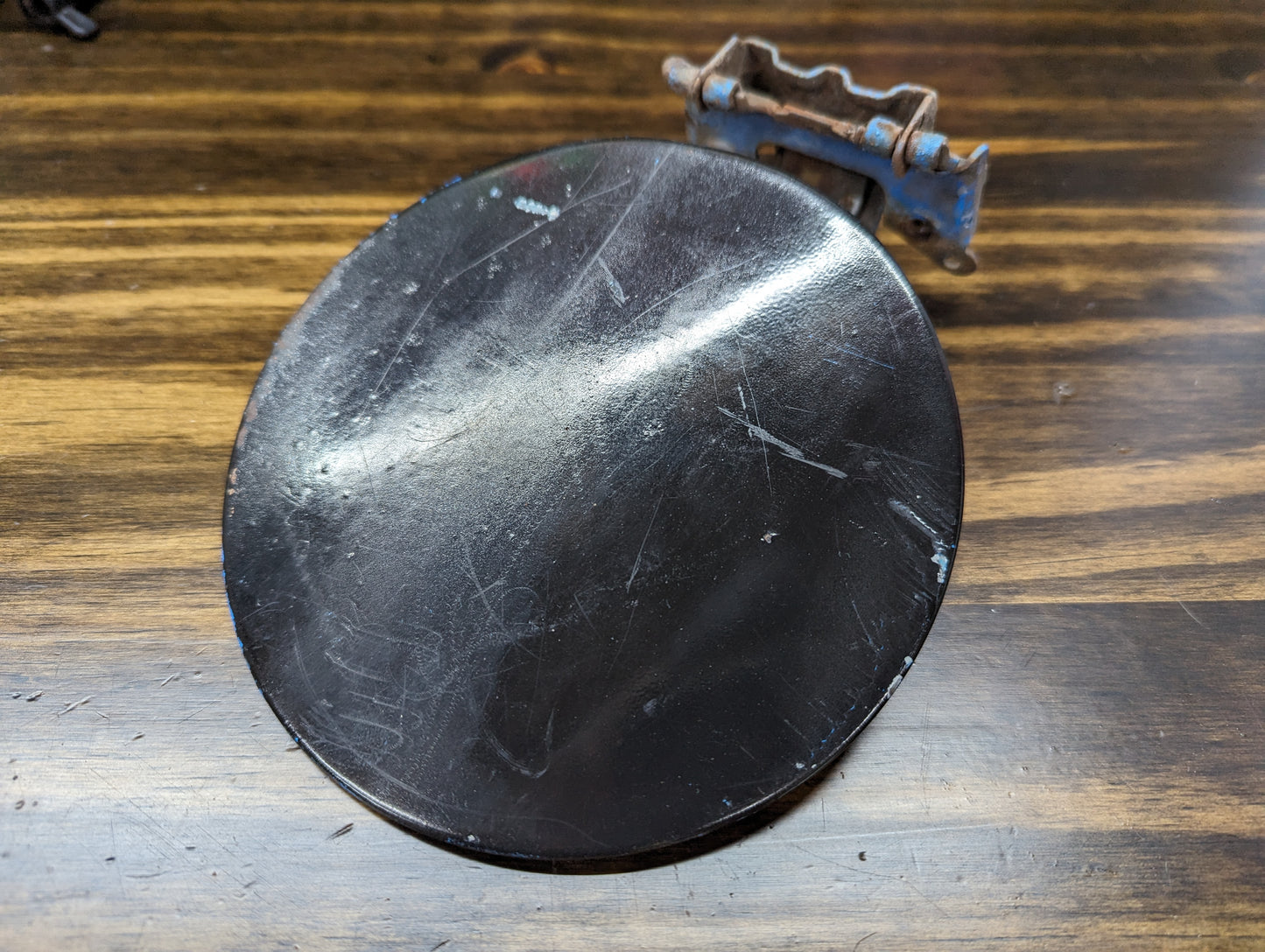 A scratched, round, black General Motors OEM Fuel Filler Neck Door with hinge and rusted bracket—fits 2001-2004 Chevy S10 & GMC Sonoma—resting on a brown wooden surface.