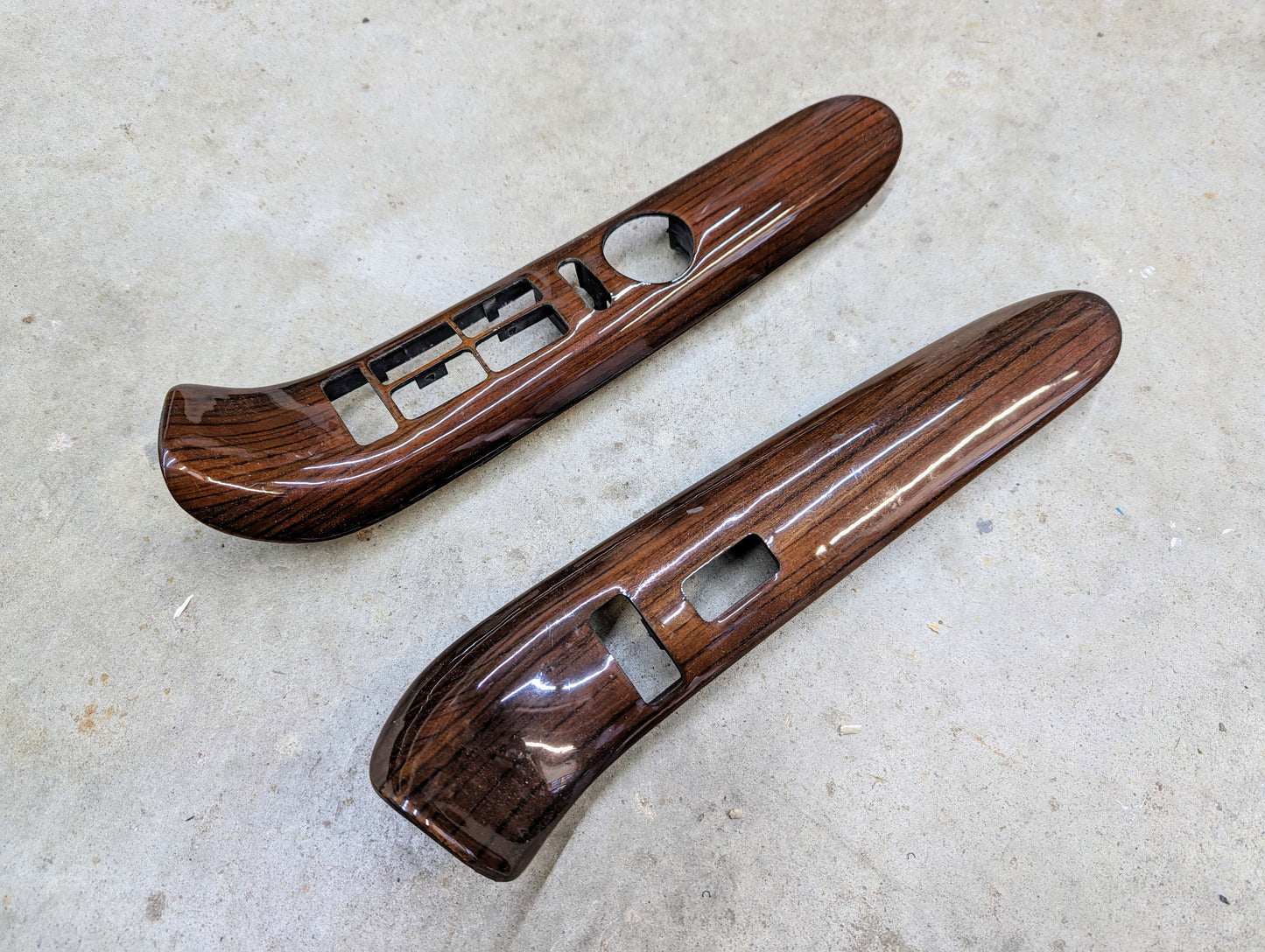A pair of General Motors OEM woodgrain power switch bezels (LH & RH) for 1999-2000 Escalade/Denali, featuring switch cutouts, shown on a light concrete surface.
