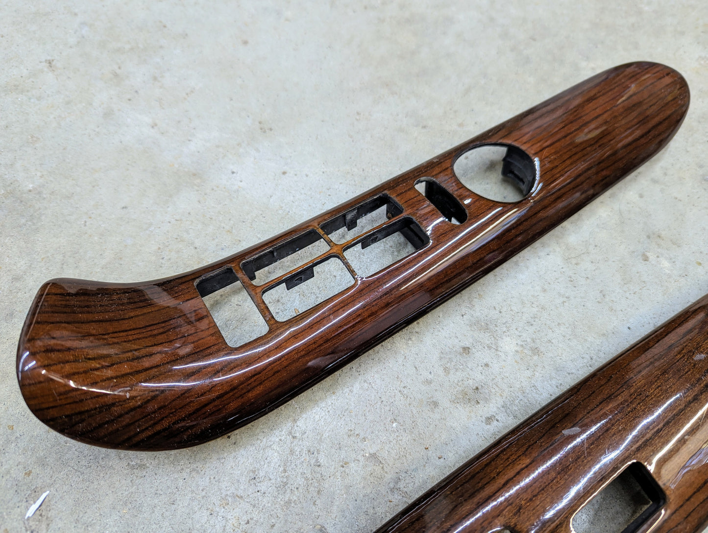A General Motors OEM Woodgrain Pair LH RH Power Switch Bezel for 1999-2000 Escalade Denali is shown on a light, textured surface. Features cutouts for window and lock controls and fits various GMC models.