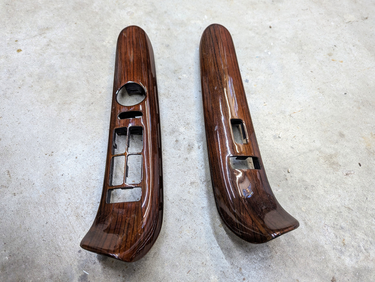 The General Motors OEM Woodgrain Pair LH RH Power Switch Bezels for 1999-2000 Escalade Denali are shown side by side on a light gray concrete surface, ideal for upgrading your interior with an authentic woodgrain finish.