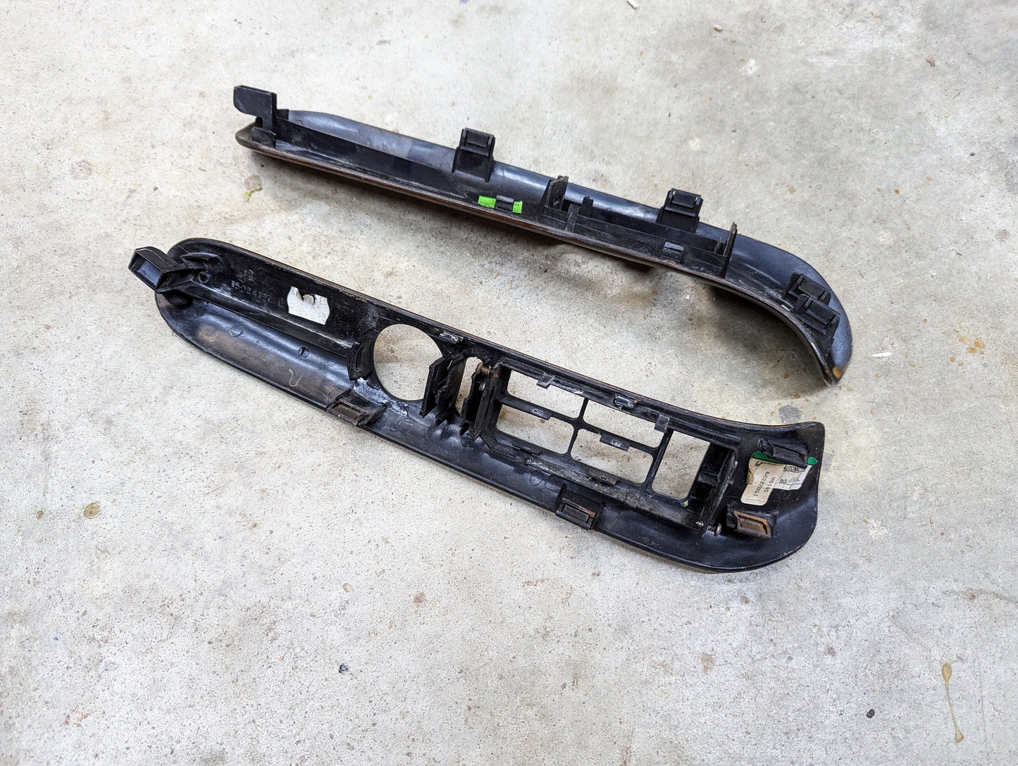 Two OEM woodgrain power switch bezels by General Motors for 1999–2000 Escalade and Denali models are shown lying on a concrete floor; both used parts display visible clips, mounting points, and matching cutouts.