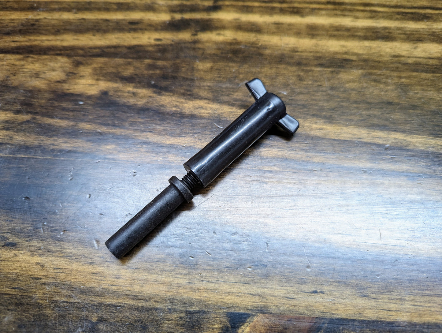 A General Motors NOS Cold Air Intake Hat Hold Down Wing Nut & Stud for 4.3/5.0/5.7L 1996–2007 GM Trucks rests on a wooden surface, its worn threaded shaft and T-handle showing signs of frequent use.