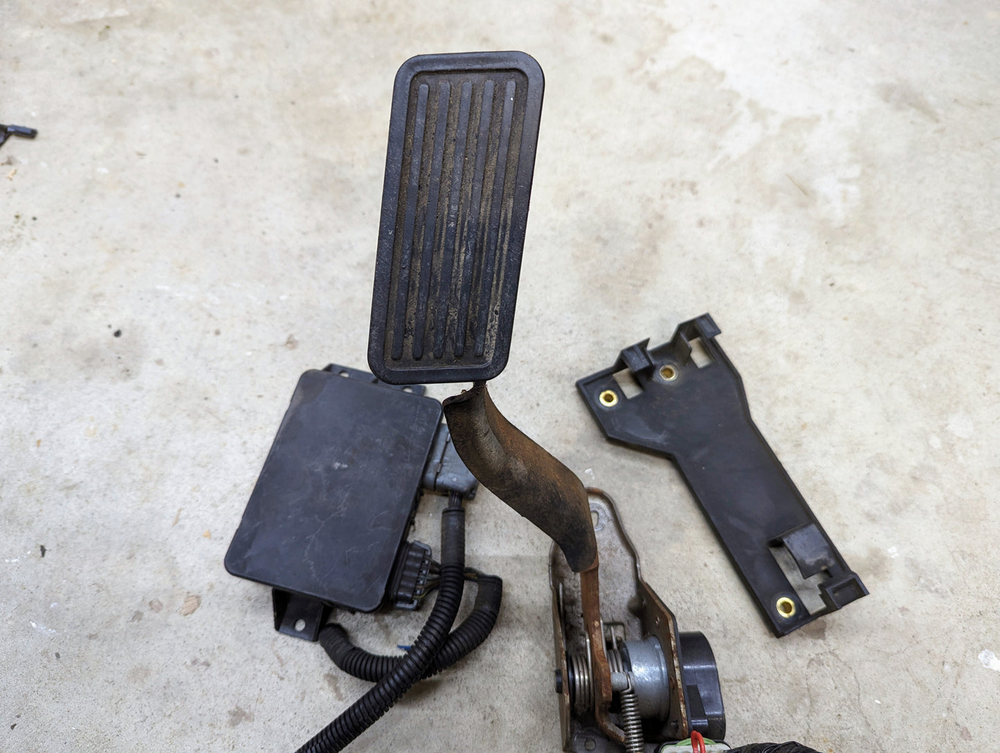 A close-up of a General Motors LS Drive By Wire Throttle Pedal & TAC Module w/Bracket for 2000–2002 Chevy Silverado, Tahoe, GMC Yukon, and more, shown with wiring on concrete beside a detached black plastic cover.