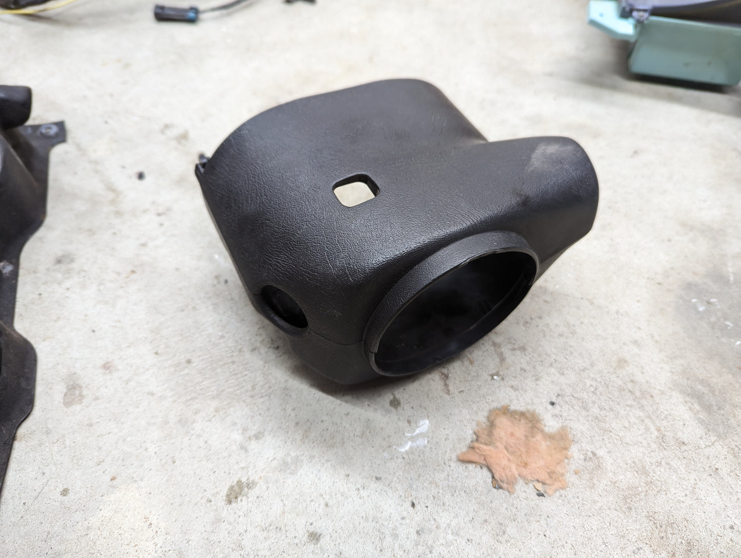 A black General Motors OEM Steering Column Cover Set for 1998-2005 Chevy S10 and Blazer rests on concrete, featuring rectangular and circular openings. Small parts and a dried leaf are scattered nearby.