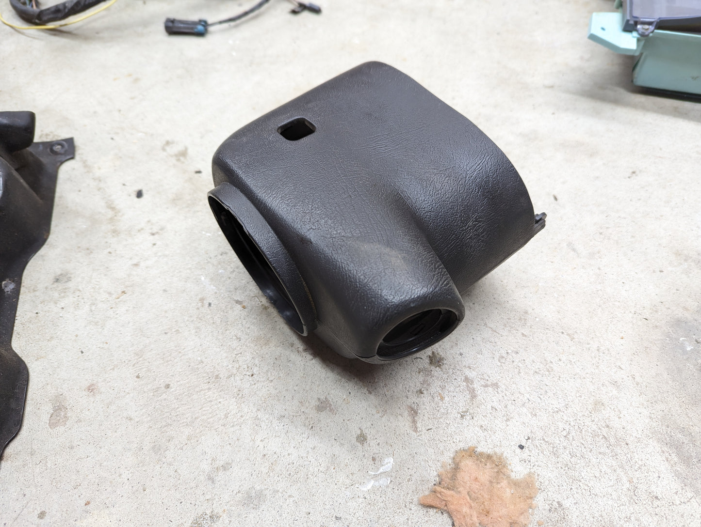 A black, textured plastic General Motors OEM Steering Column Cover Set for 1998-2005 Chevy S10, Blazer, and more rests on a concrete floor amid small debris and other car parts.
