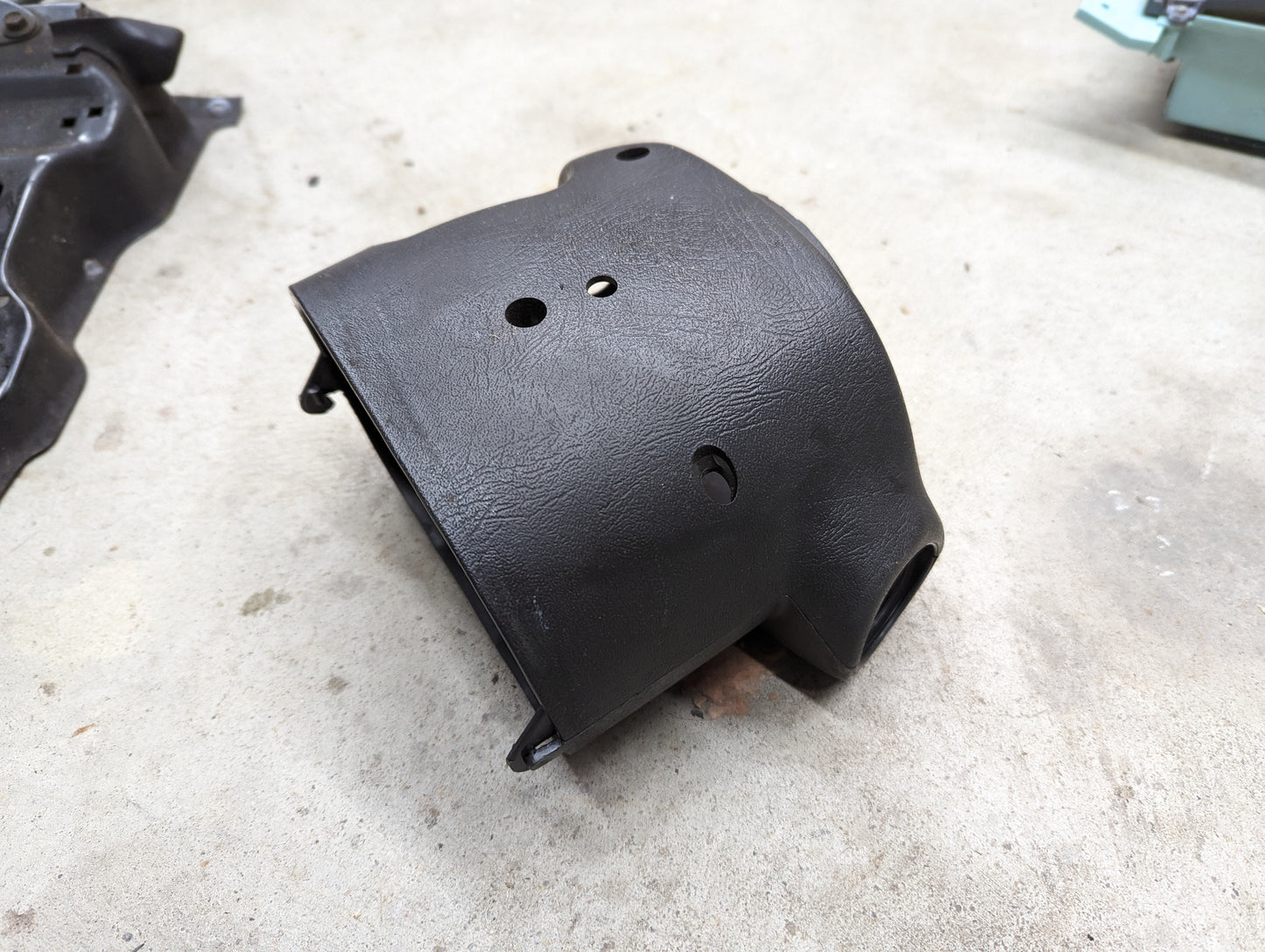 A black, textured OEM Steering Column Cover Set by General Motors for 1998-2005 Chevy S10, Blazer, and more features multiple openings, a curved cylindrical section, and mounting brackets, shown resting on a concrete floor.