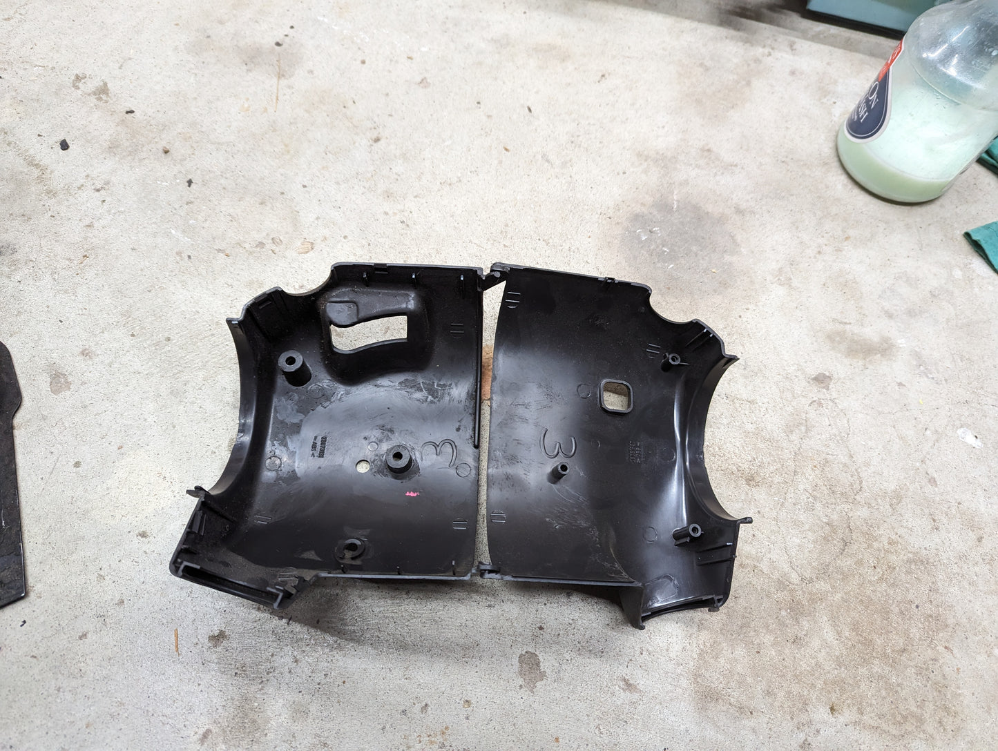 Two black plastic parts from the General Motors OEM Steering Column Cover Set for 1998-2005 Chevy S10, Blazer, and more are shown on a concrete surface. A green and white bottle and green cloth are visible in the upper right corner.