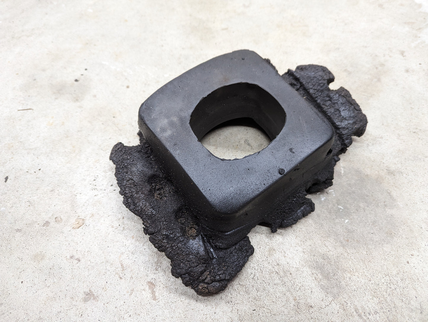 A rugged, black OEM Manual Floor Shifter Insulator Foam by General Motors for 1998-2005 Chevy S10, Blazer, and more features a square center hole and rests on textured concrete with unevenly fused material at its base.