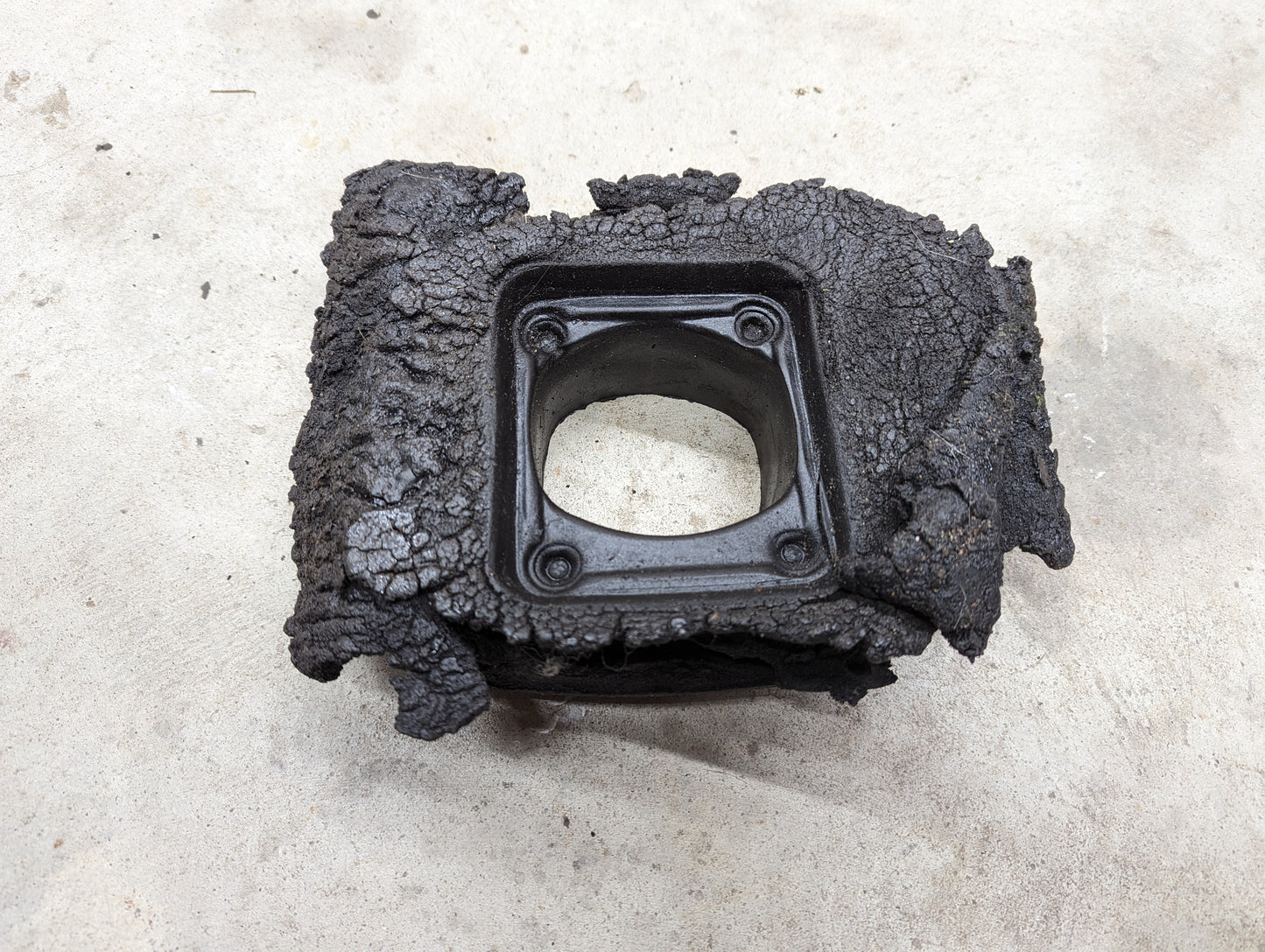 A black, rectangular General Motors OEM Manual Floor Shifter Insulator Foam for 1998–2005 Chevy S10 and Blazer with a hollow square center and four bolt holes rests on rough concrete, showing signs of damage.