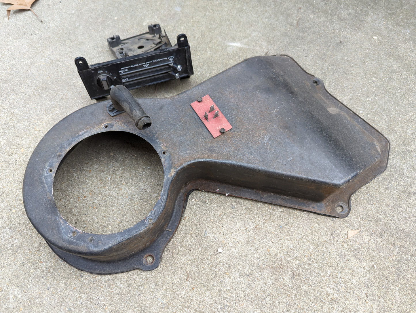 An OEM A/C Delete Heater Box & Control Panel by General Motors for 1982-1985 S10, Blazer, and more—including the metal fan shroud, bracket, and black component—lie on a concrete surface.