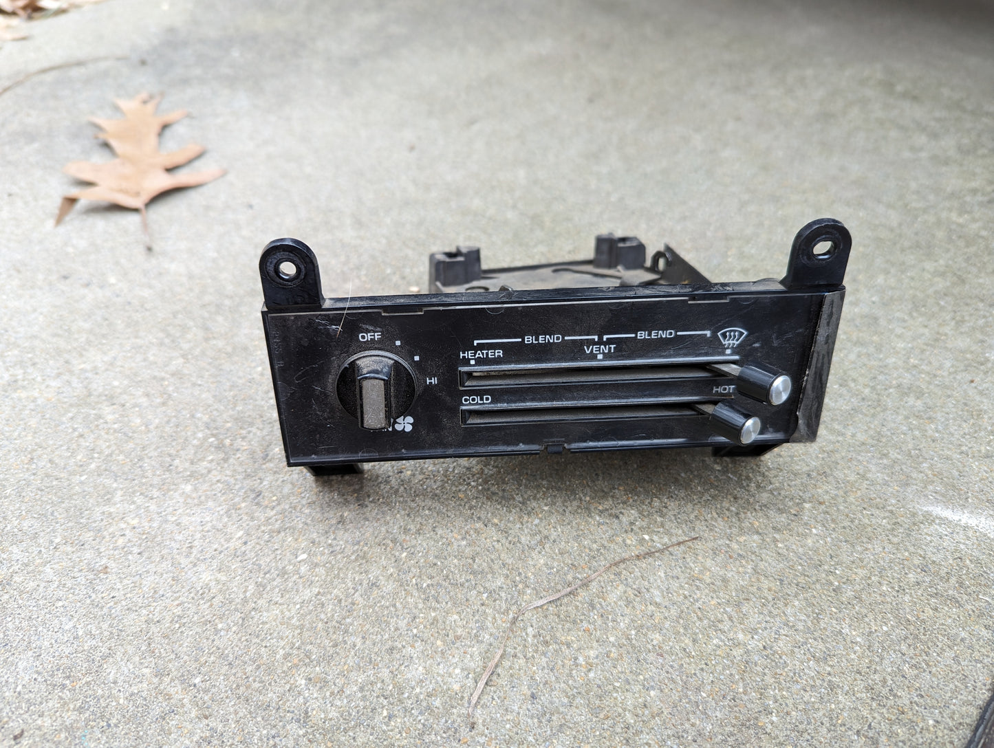 A General Motors OEM A/C Delete Heater Box & Control Panel for 1982-1985 S10, Blazer, and more rests on concrete. Sliders for heater, vent, and temperature are visible; a nearby brown leaf hints at possible modifications.
