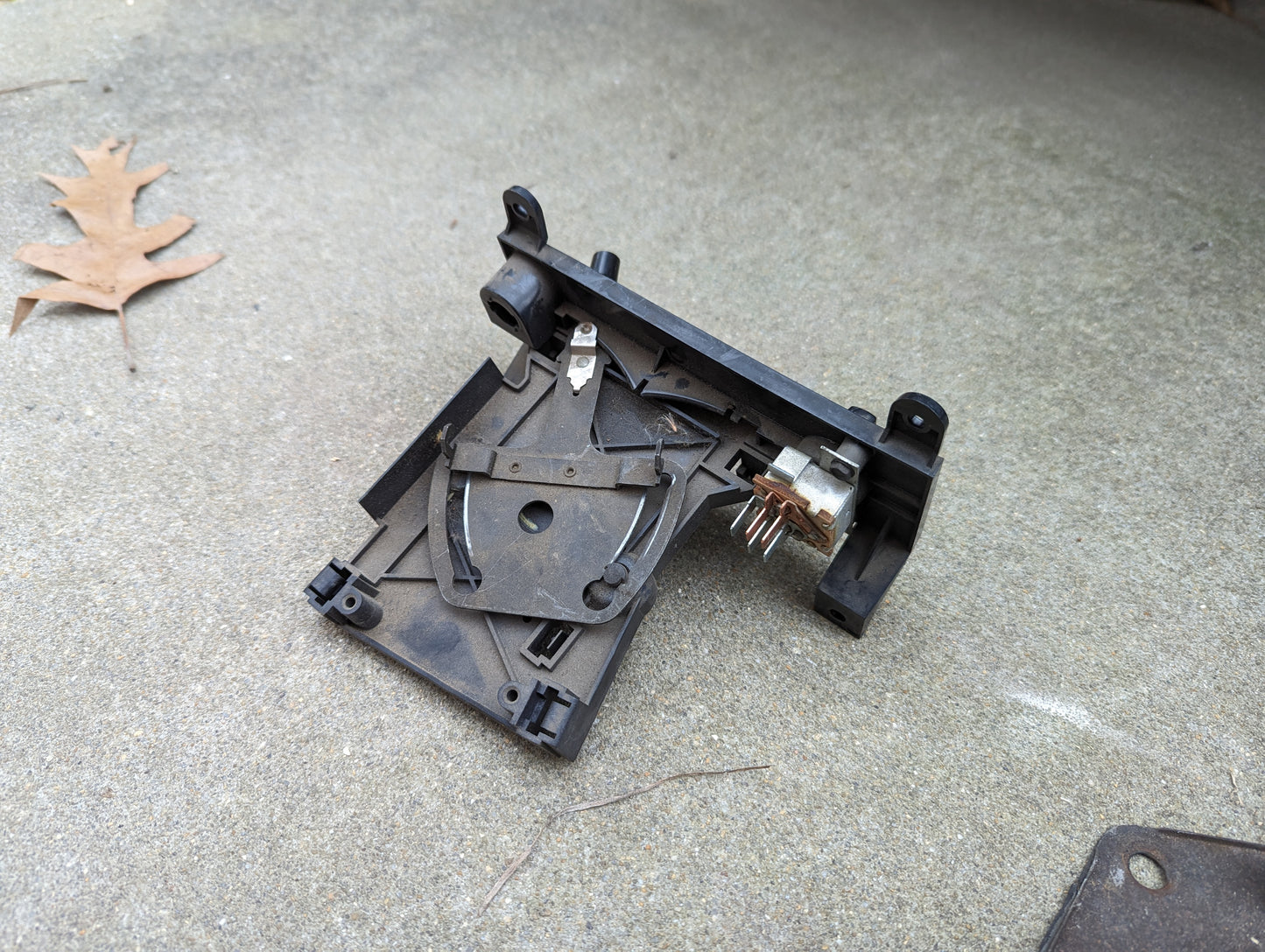 A close-up of a disassembled General Motors OEM A/C Delete Heater Box & Control Panel for 1982-1985 S10, Blazer, and more, showing exposed metal pins on concrete beside a dry oak leaf.
