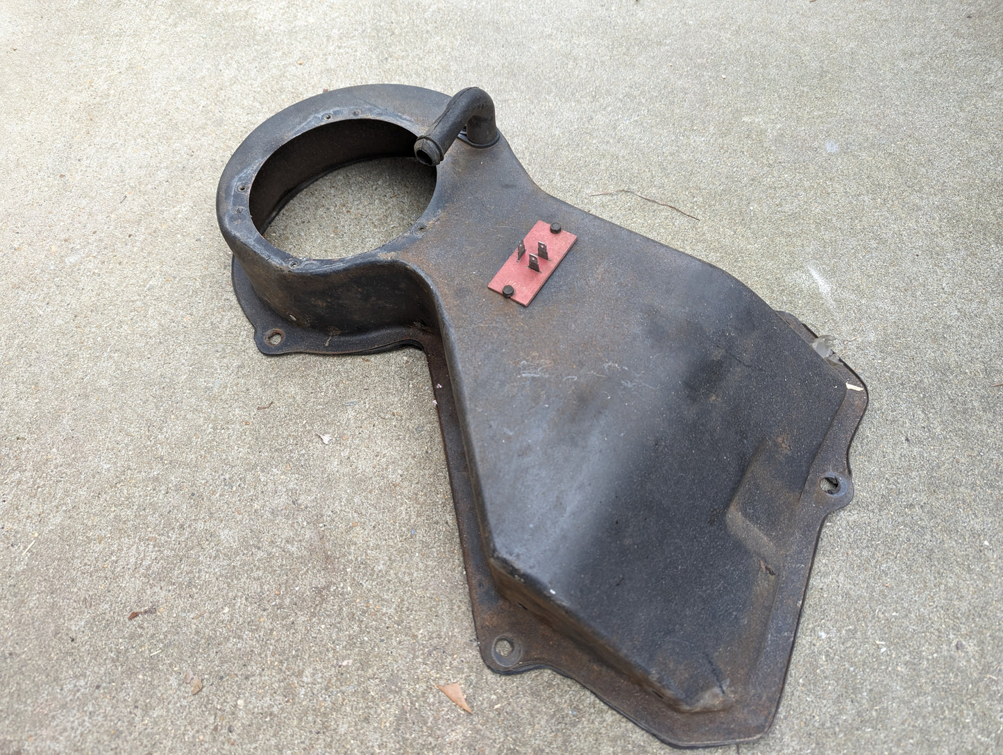 A worn OEM A/C Delete Heater Box & Control Panel by General Motors for 1982-1985 S10, Blazer, and more shows rust and wear on a concrete surface. This black metal part features a large circular opening and a small red rectangular piece.