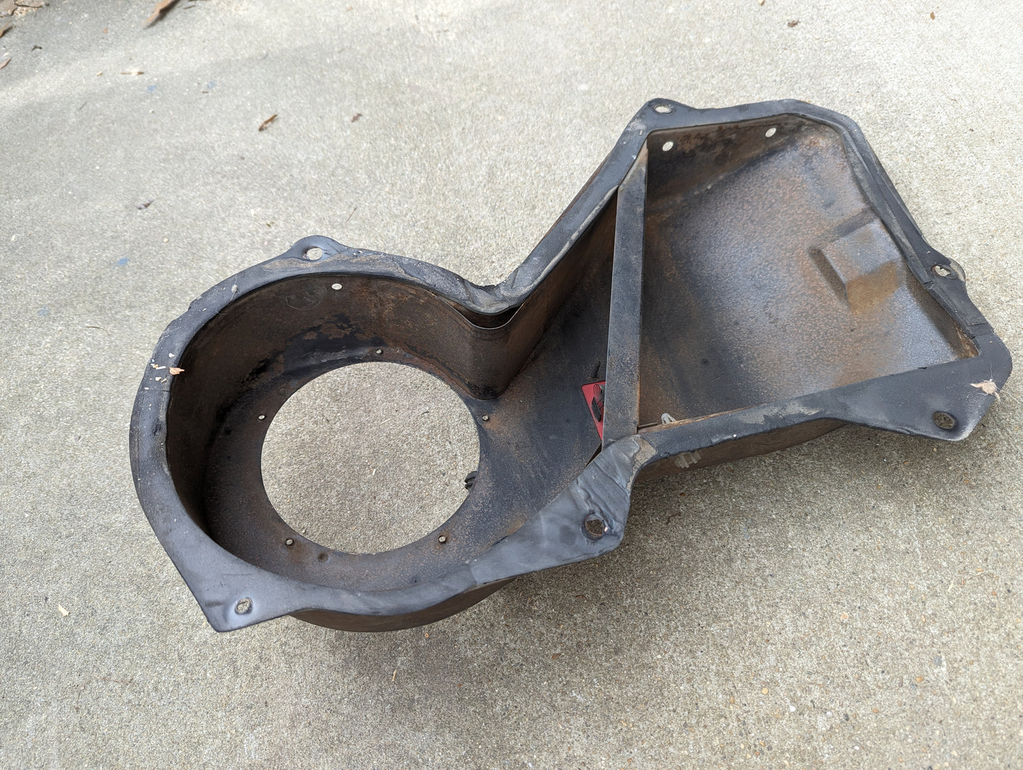 A worn OEM A/C Delete Heater Box & Control Panel by General Motors for 1982-1985 S10, Blazer, and more—with a large circular opening and several bolt holes—rests on concrete, showing signs of age.