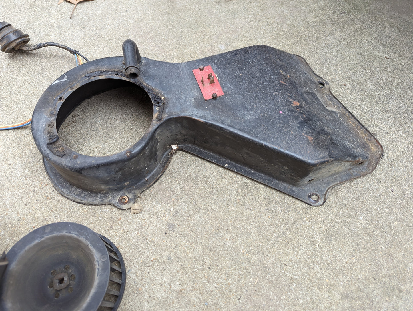 A used General Motors OEM A/C delete heater box and control panel swap part for 1986-1994 S10, Blazer, and more sits on concrete with a round opening, red mounting clip, attached wires, and a separated fan blade.