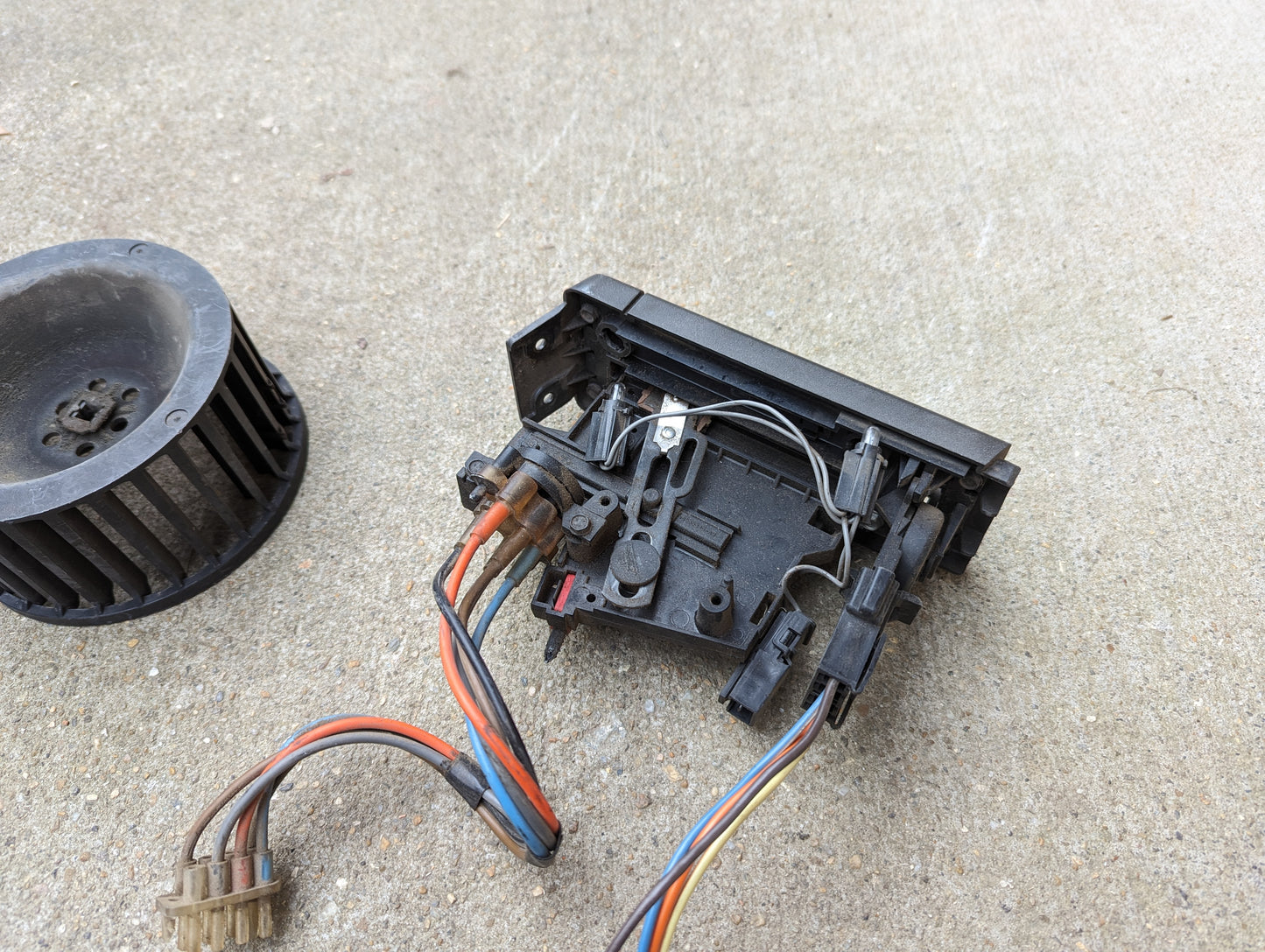 A small electrical component from the General Motors OEM A/C Delete Heater Box & Control Panel Swap Parts for 1986-1994 S10, Blazer, and more—with multicolored wires, a circuit board, and a plastic fan—rests on rough concrete.
