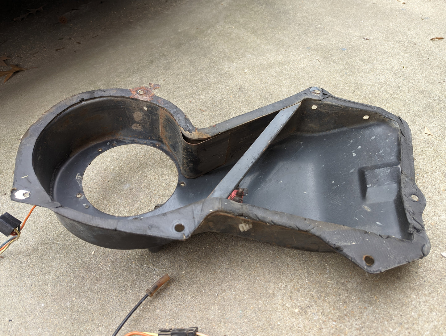 A General Motors OEM A/C Delete Heater Box & Control Panel Swap part for 1986-1994 S10, Blazer, and more, featuring a black plastic body with round and rectangular openings, is shown resting on a concrete surface amid wires and dirt.