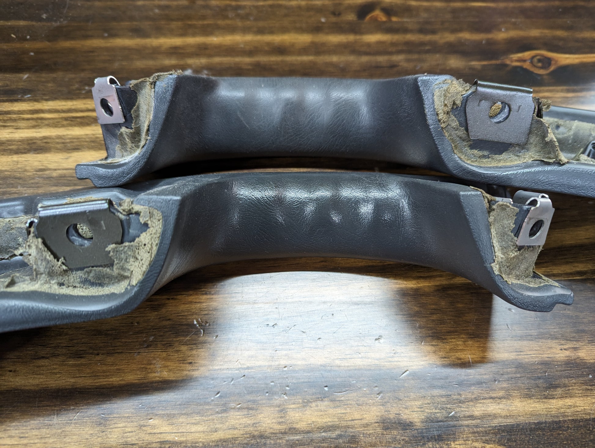 Two black padded car grab handles by The Ugly Truckling, likely KIT Door Panel Armrest Repair for 1998-2005 Chevy S10/Blazer/GMC Sonoma/Jimmy, show exposed worn foam and metal brackets at the ends, resting on a wooden surface. Signs of damage and aging are visible.