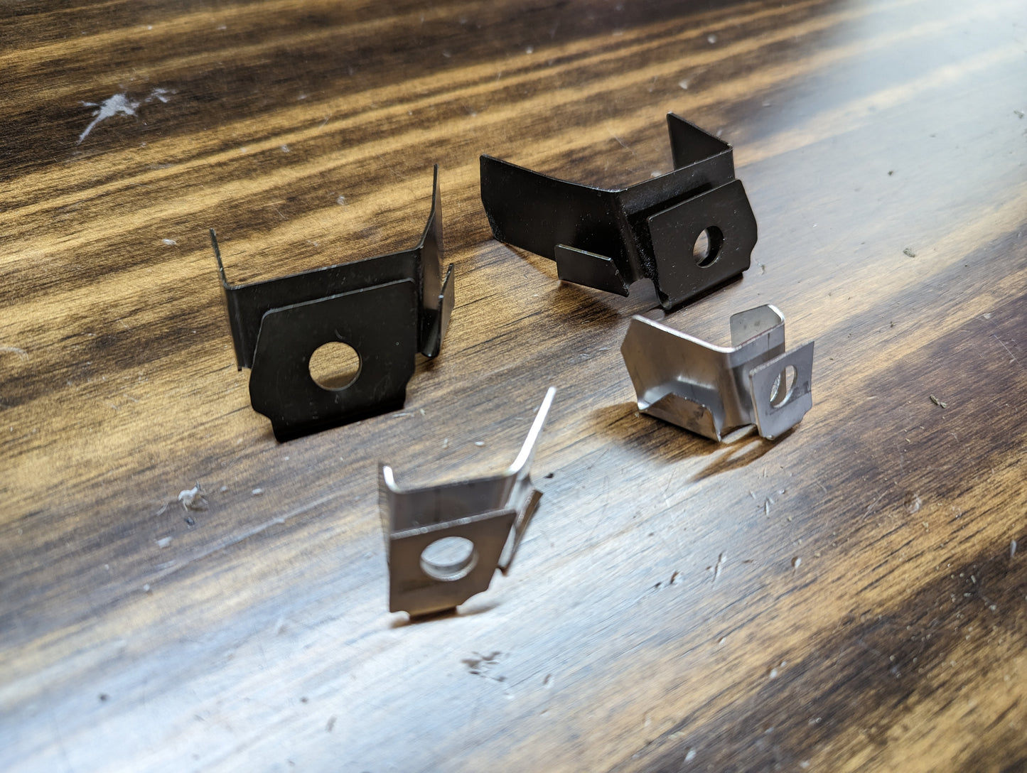 Four metal clips from The Ugly Truckling KIT Door Panel Armrest Repair for 1998-2005 Chevy S10, Blazer, GMC Sonoma, and Jimmy are arranged on wood; two are black and two are silver, each with a hole in the center.