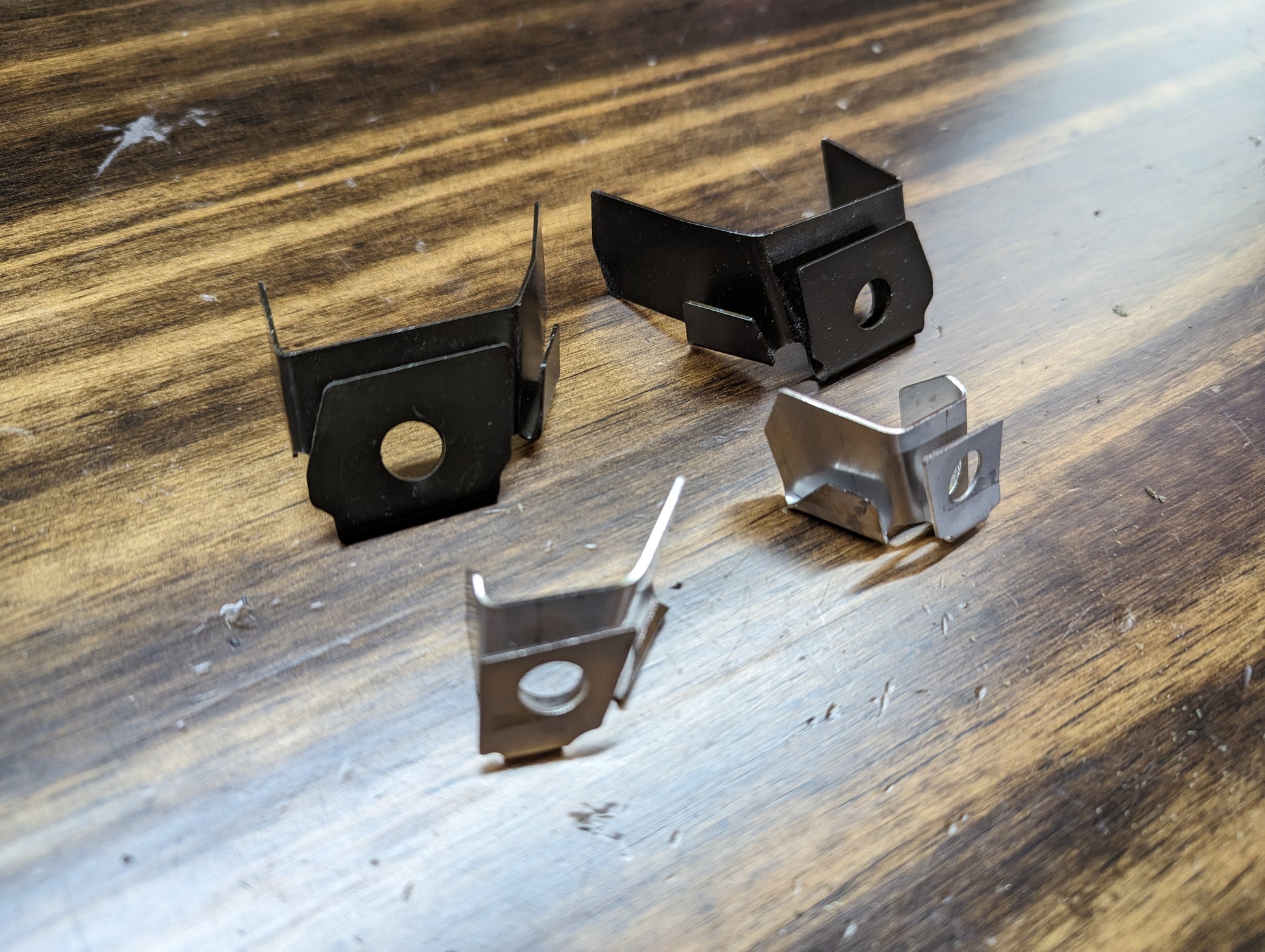 Four metal clips from The Ugly Truckling KIT Door Panel Armrest Repair for 1998-2005 Chevy S10, Blazer, GMC Sonoma, and Jimmy are arranged on wood; two are black and two are silver, each with a hole in the center.