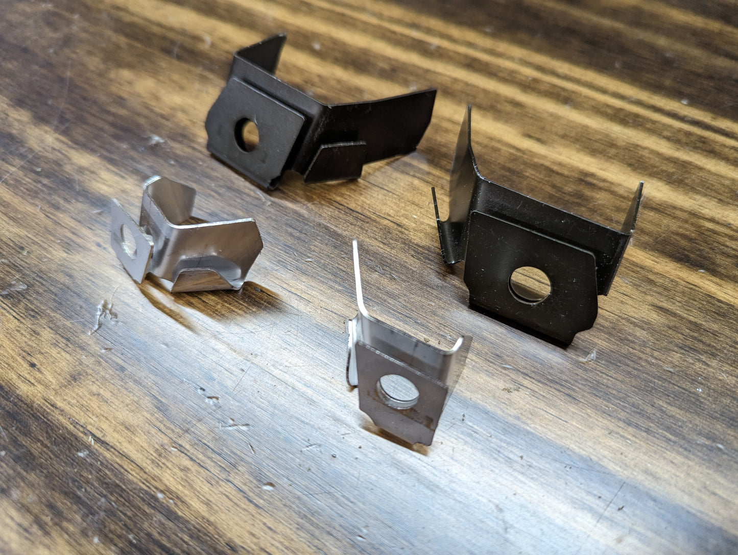Four small metal brackets from The Ugly Truckling KIT Door Panel Armrest Repair for 1998-2005 Chevy S10, Blazer, GMC Sonoma, and Jimmy are shown on wood—two black and two silver, each with bent tabs and angled shapes.